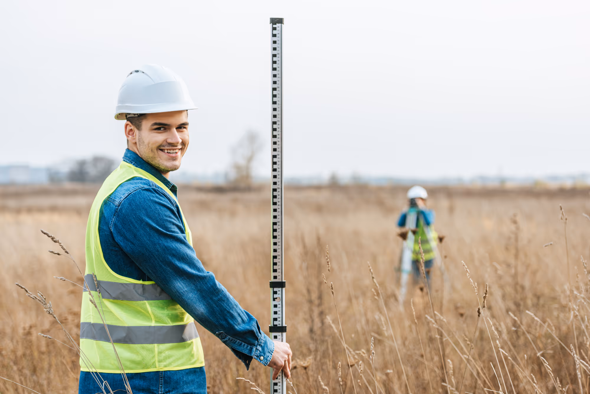 Smiling surveyor in a hard hat and reflective vest holding a leveling rod in a dry grassy field.