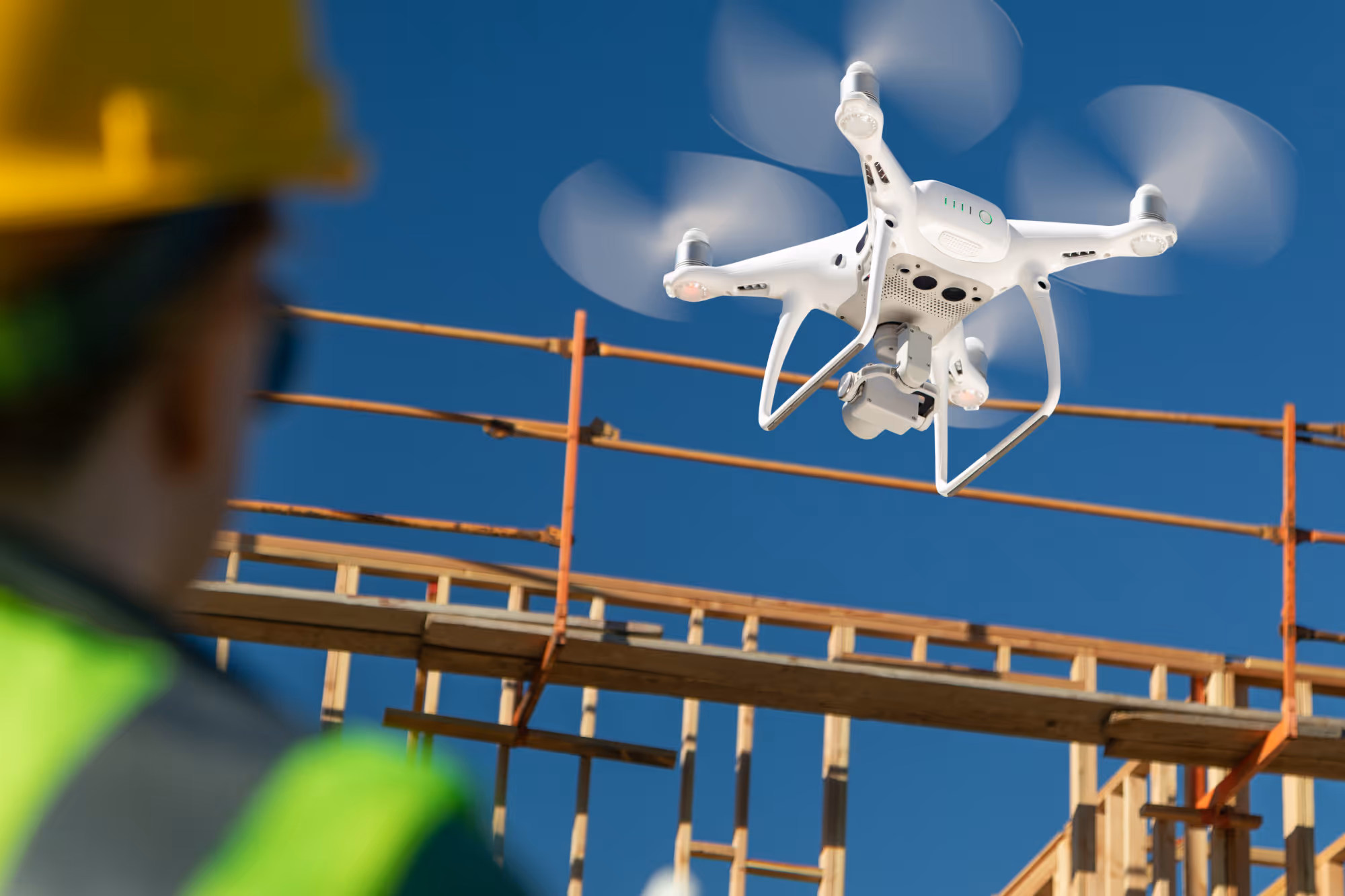 White drone flying near a construction site scaffold with a worker in a yellow helmet in foreground.