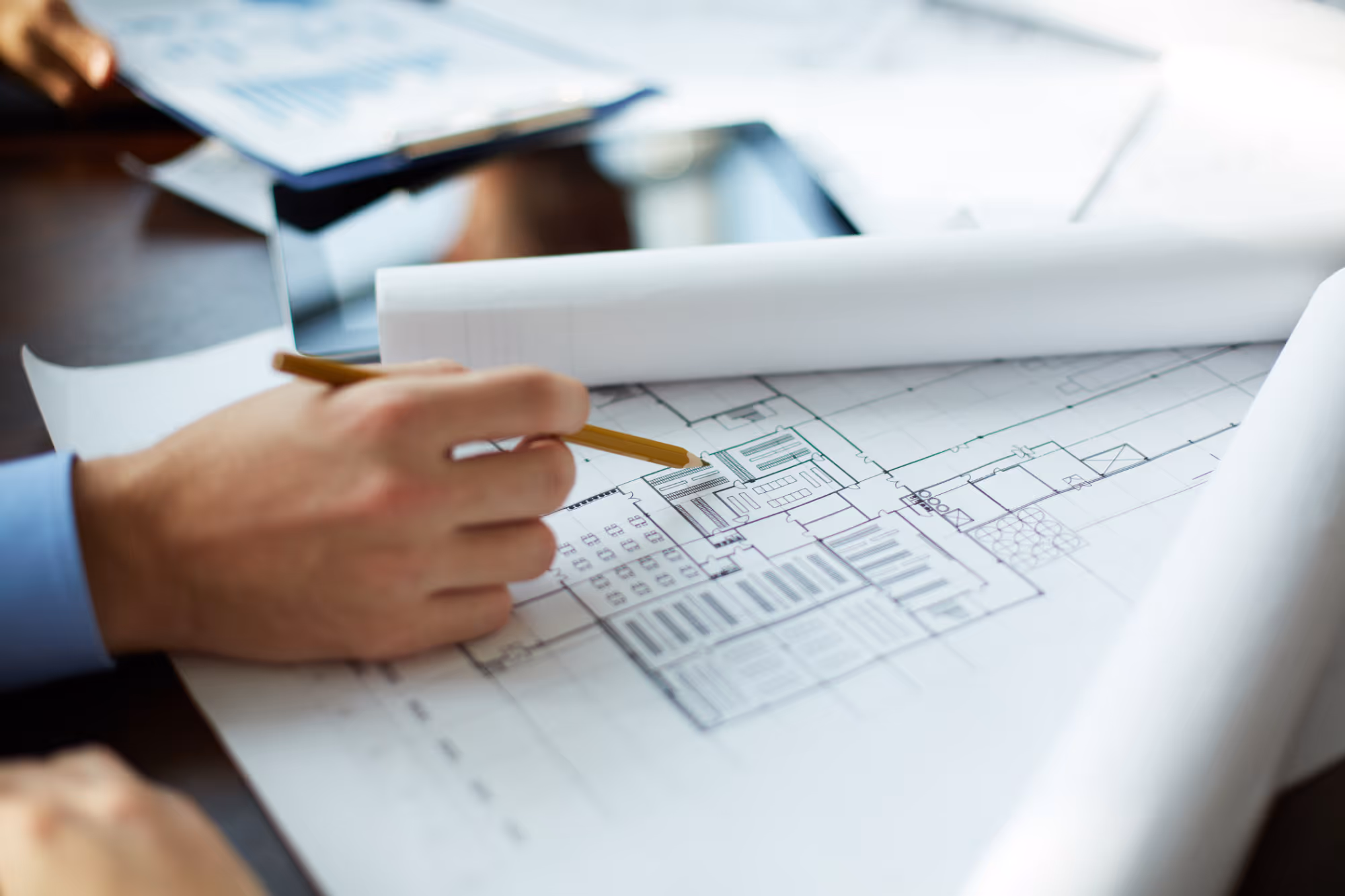Hand holding pencil pointing to architectural blueprint with tablet and papers nearby.
