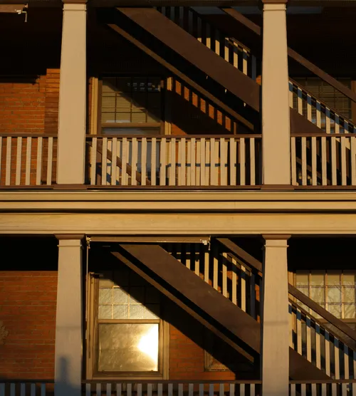 Exterior view of a brick building with white railings and dark brown fire escape stairs casting shadows in sunlight.