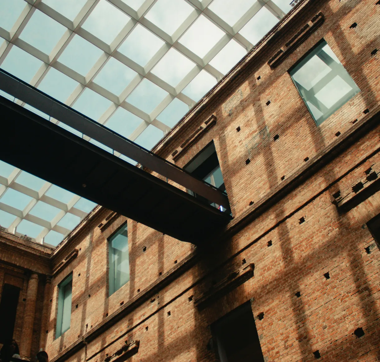 Interior view of a brick building with large windows and a black metal walkway under a glass grid ceiling.