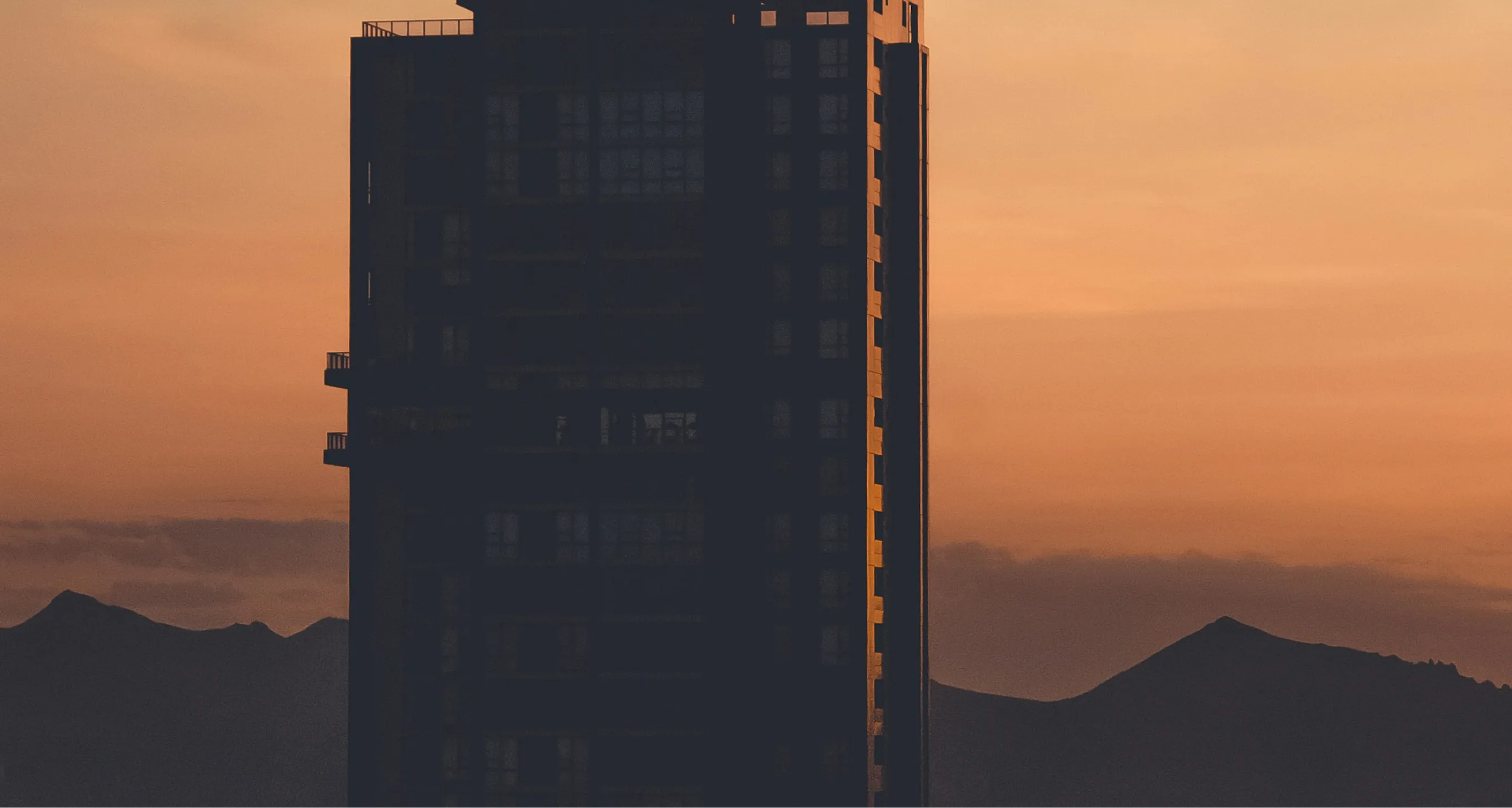 Silhouette of a tall building with mountain peaks in the background at orange sunset.