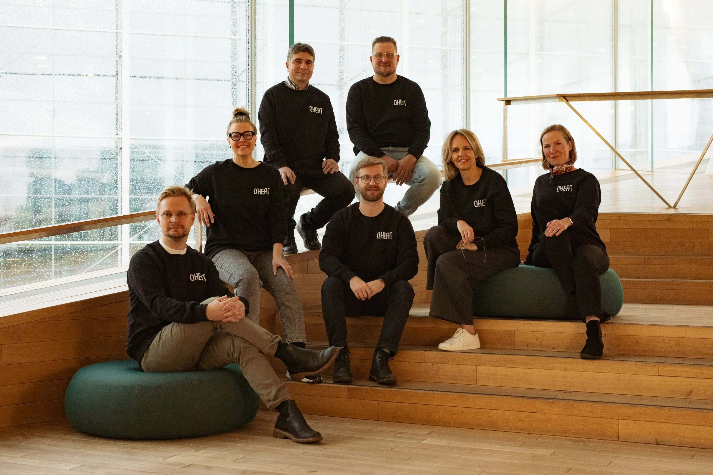 Group of seven people wearing black shirts with 'QHEAT' logo sitting on wooden steps and round cushions in a bright modern room.