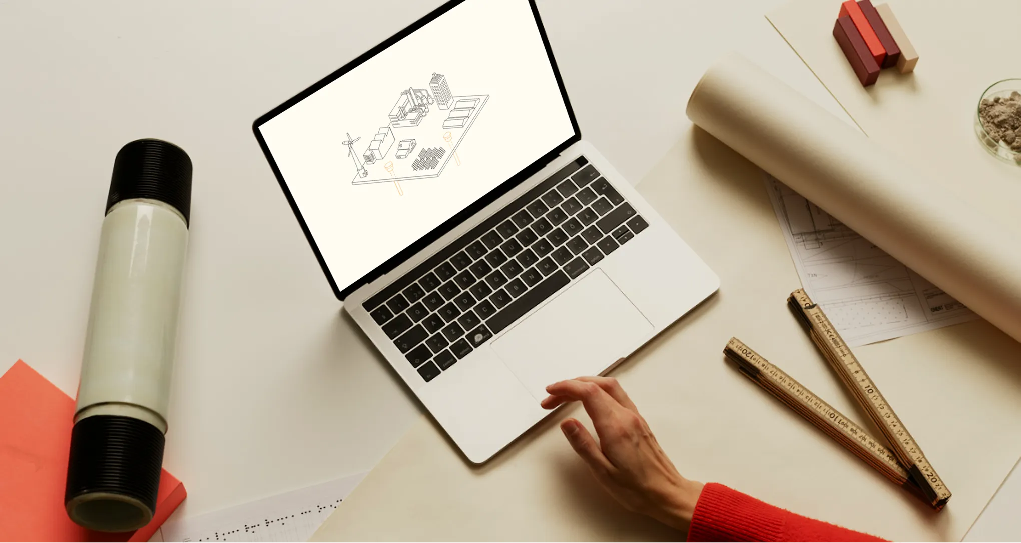 Person wearing a red sweater using a laptop showing an industrial and renewable energy schematic, surrounded by rolled-up blueprints, rulers, and design materials on a beige surface.