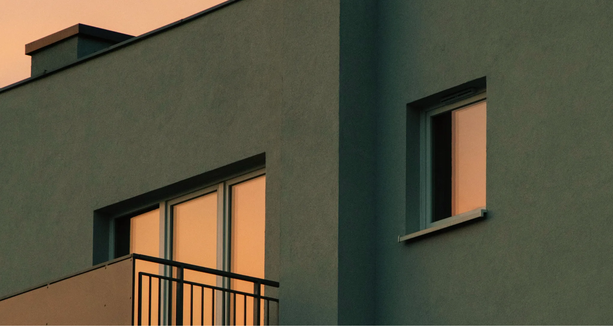 Modern building facade with two windows reflecting an orange sunset light and a balcony with a black metal railing.