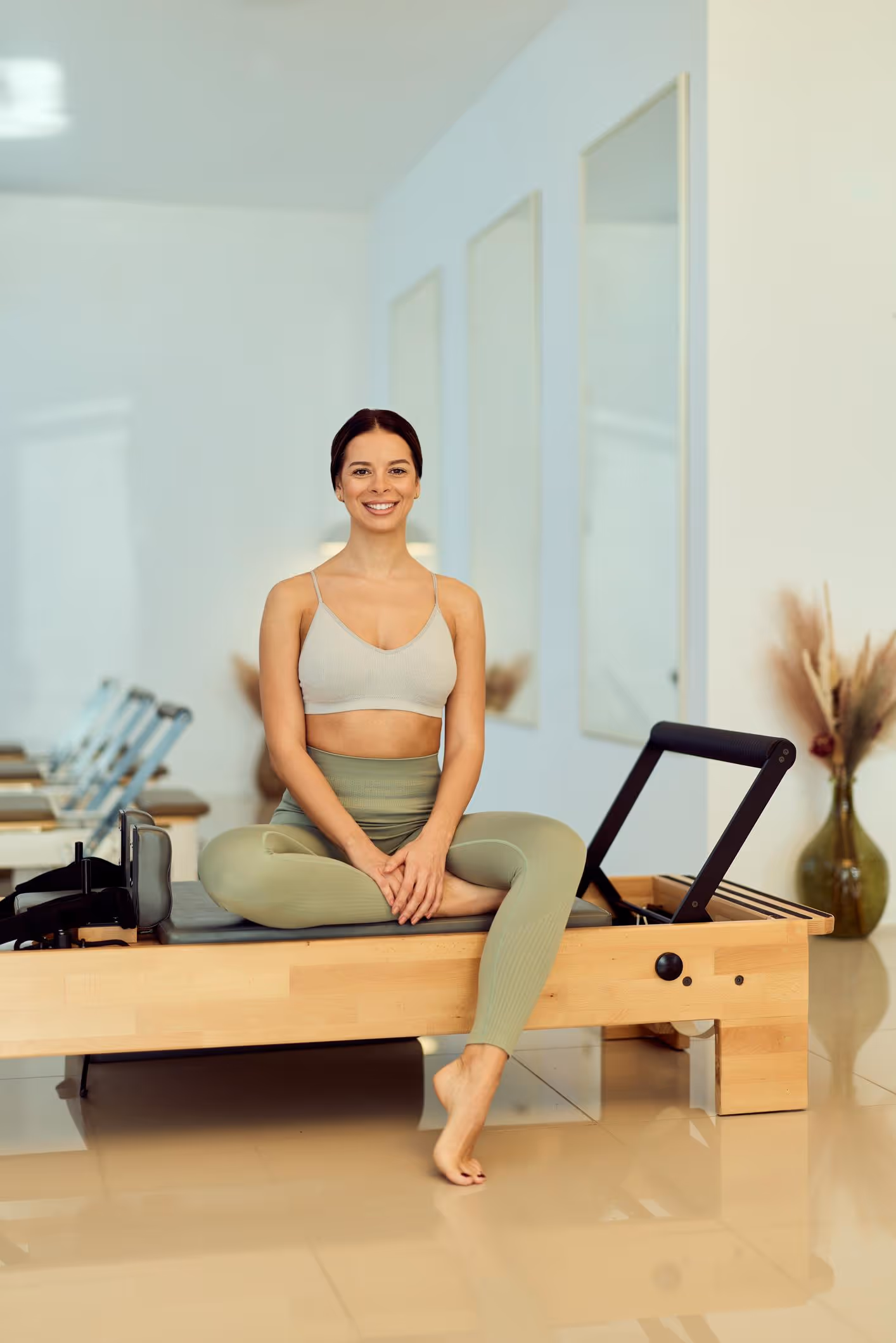 Smiling woman in workout clothes sitting cross-legged on a Pilates reformer machine indoors.