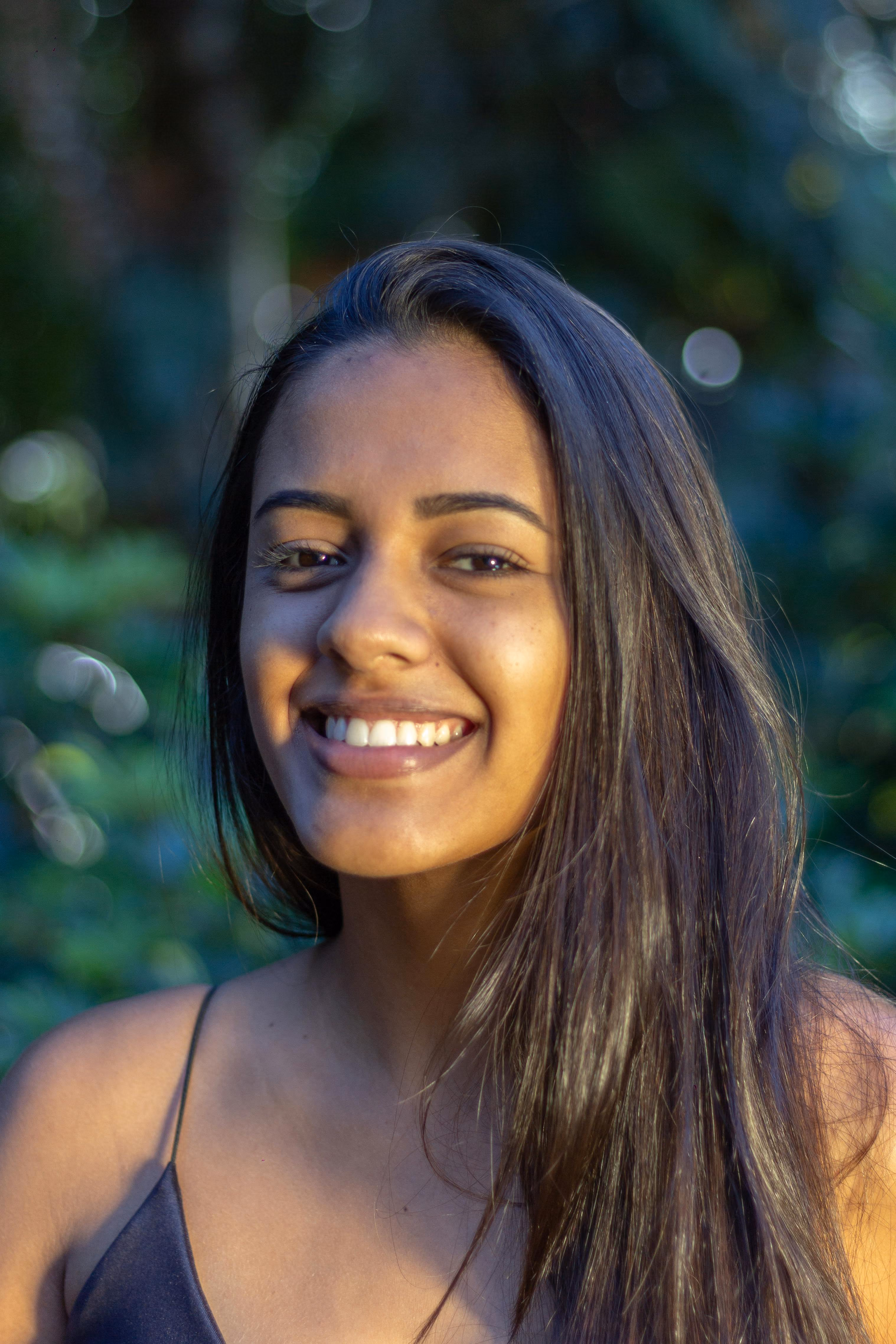 Smiling woman with long dark hair wearing a black top in an outdoor garden setting.