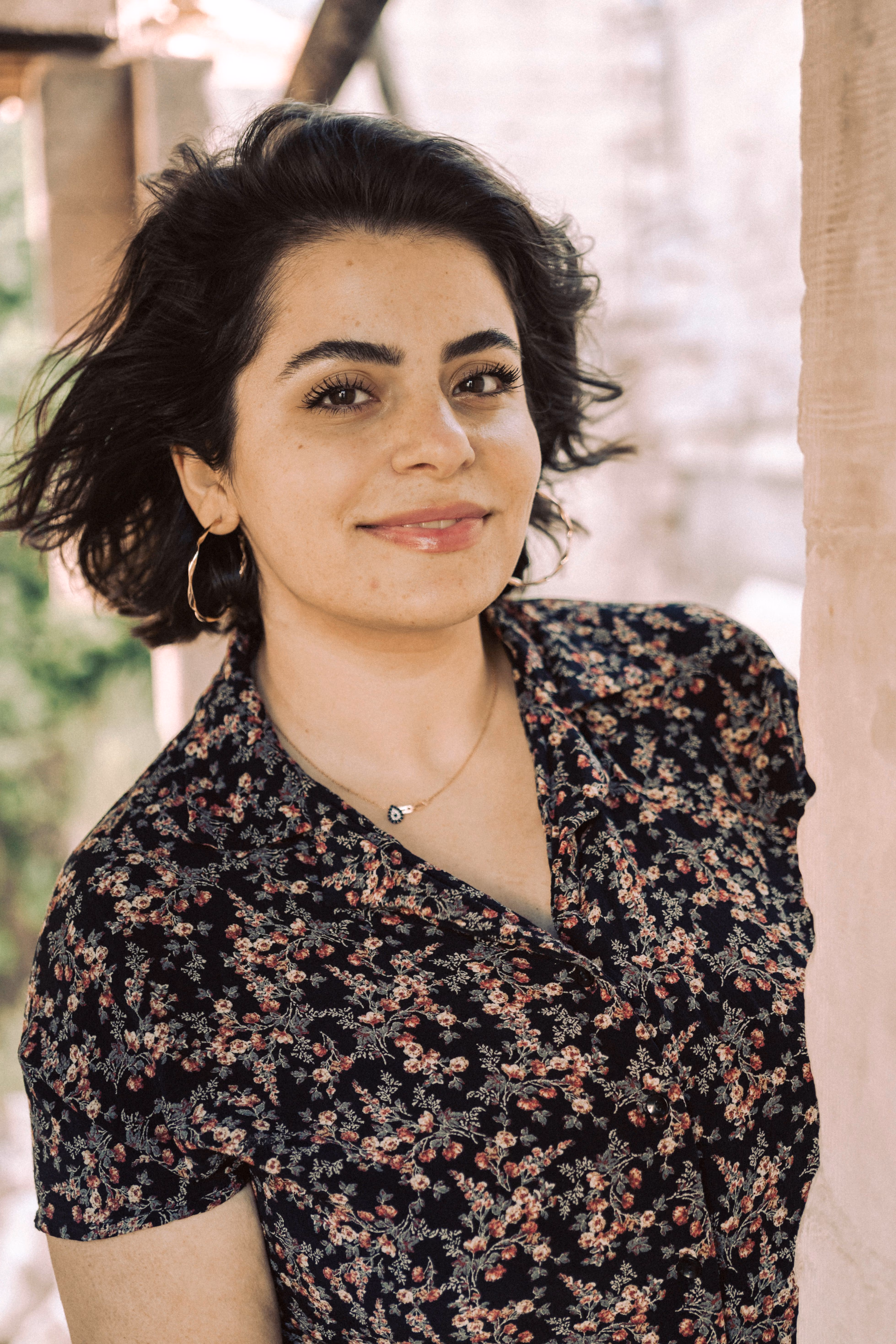 Woman with short dark hair wearing a floral blouse and gold hoop earrings, smiling softly outdoors.