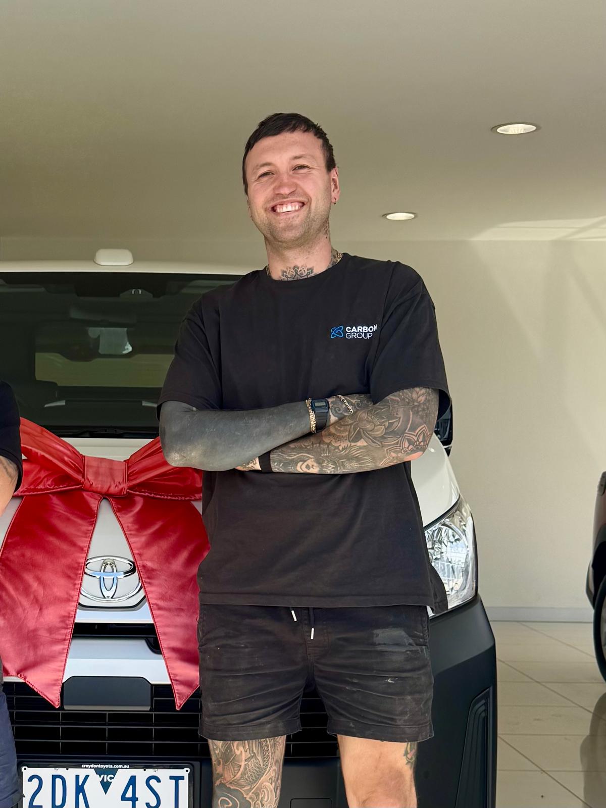 Smiling man with tattoos wearing a black Carbon Group shirt standing in front of a white vehicle with a large red bow.
