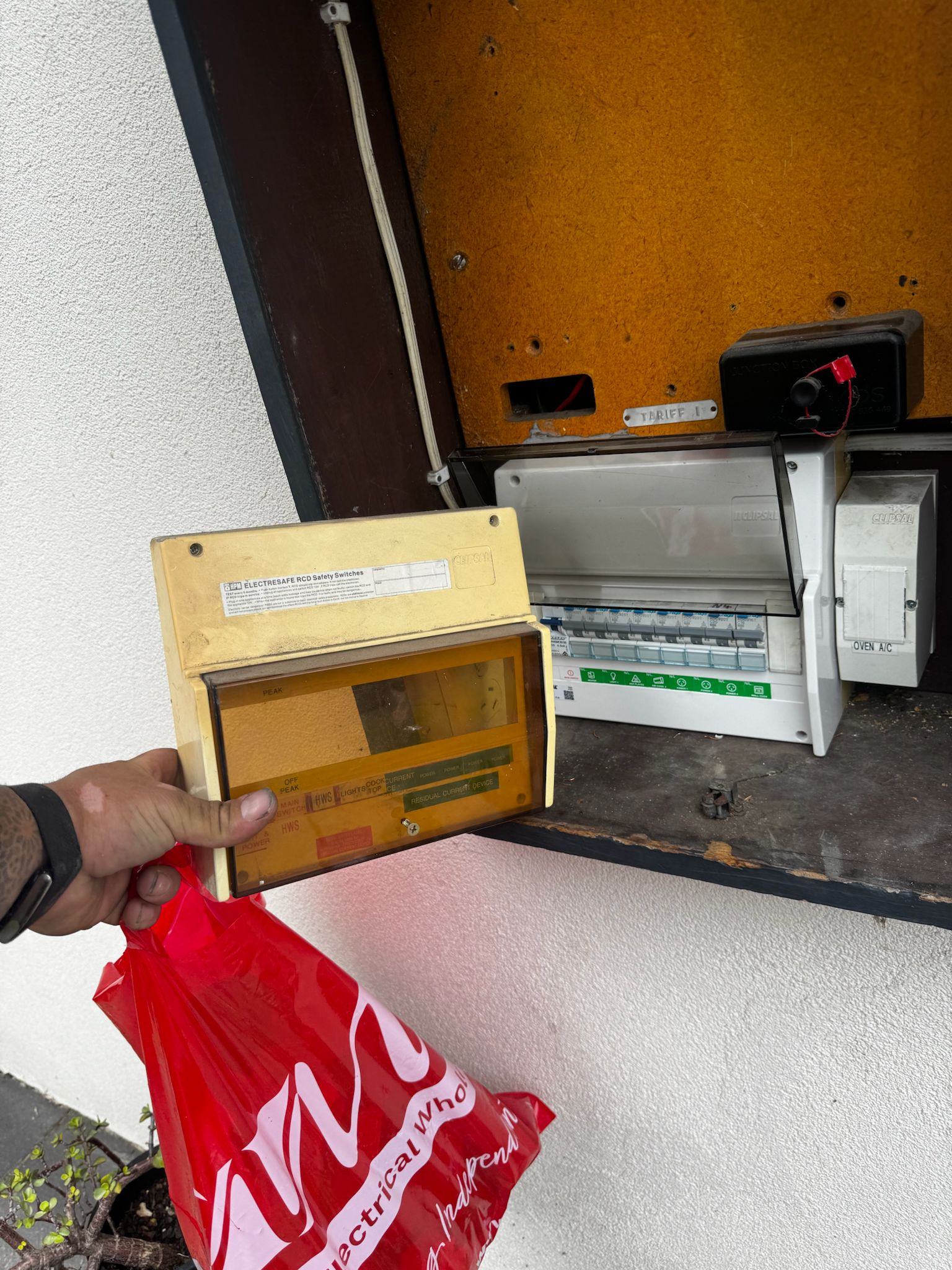 A hand holding a yellow electrical safety switch box near an open electrical panel on a wall, with a red plastic bag hanging from the wrist.