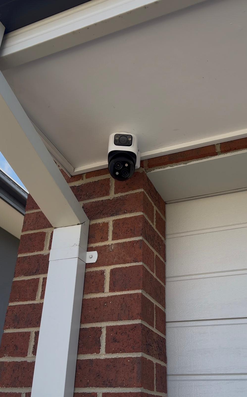 Security camera with two lenses mounted on the corner of a brick wall under a white roof overhang.