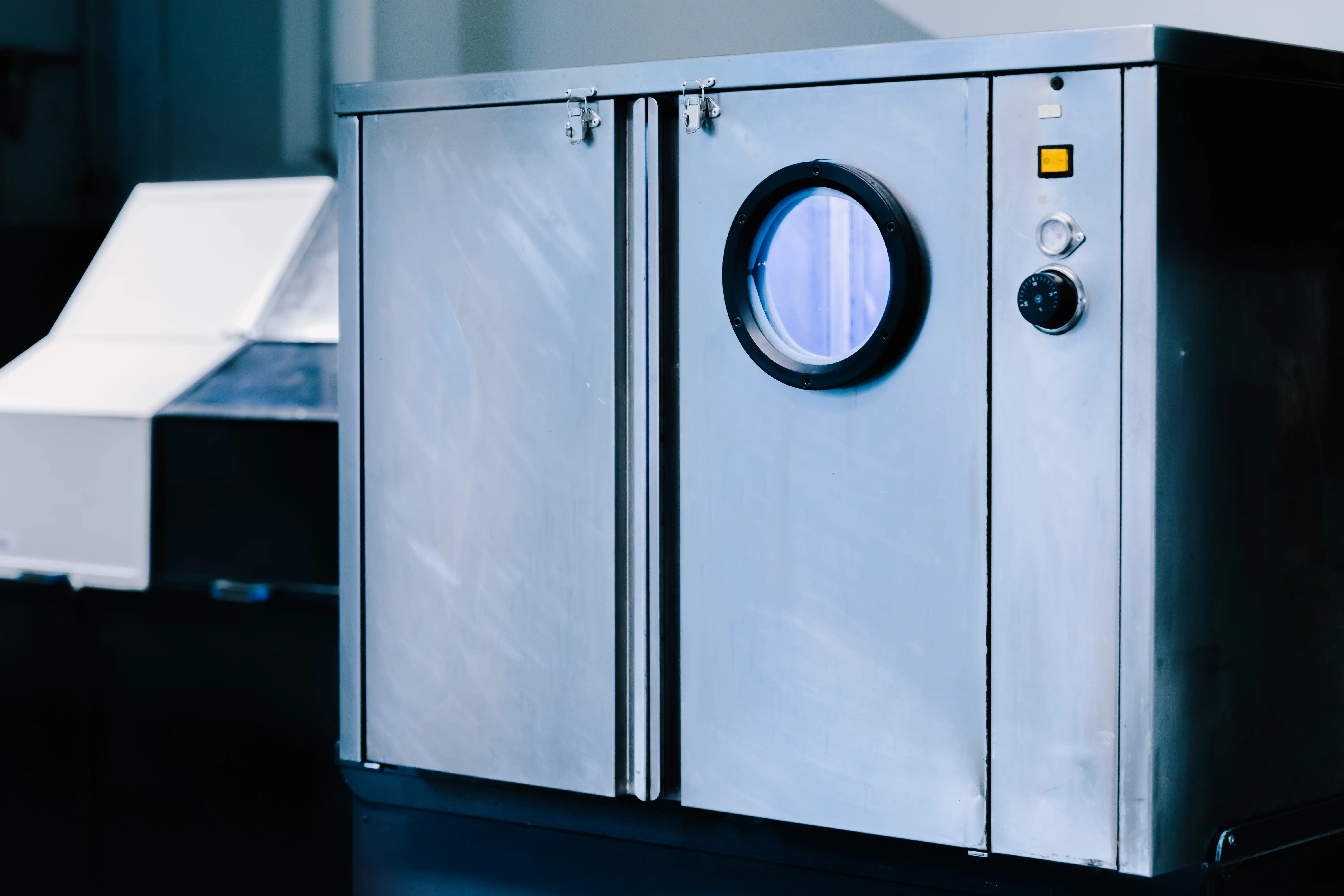 Industrial metal machine with circular window, control knob, and a yellow rectangular button.