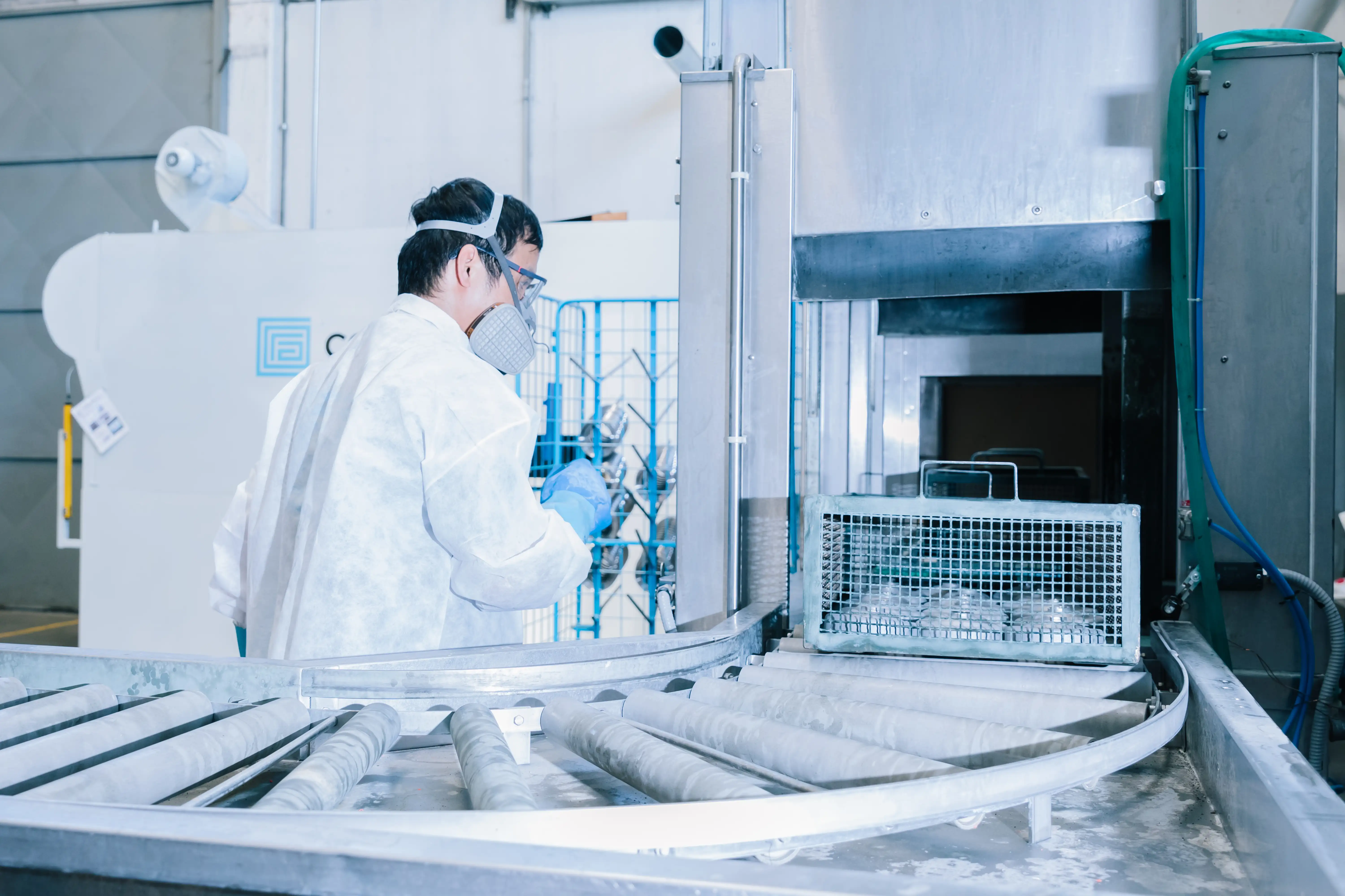 Factory worker in protective gear operating conveyor with metal mesh basket entering machine.