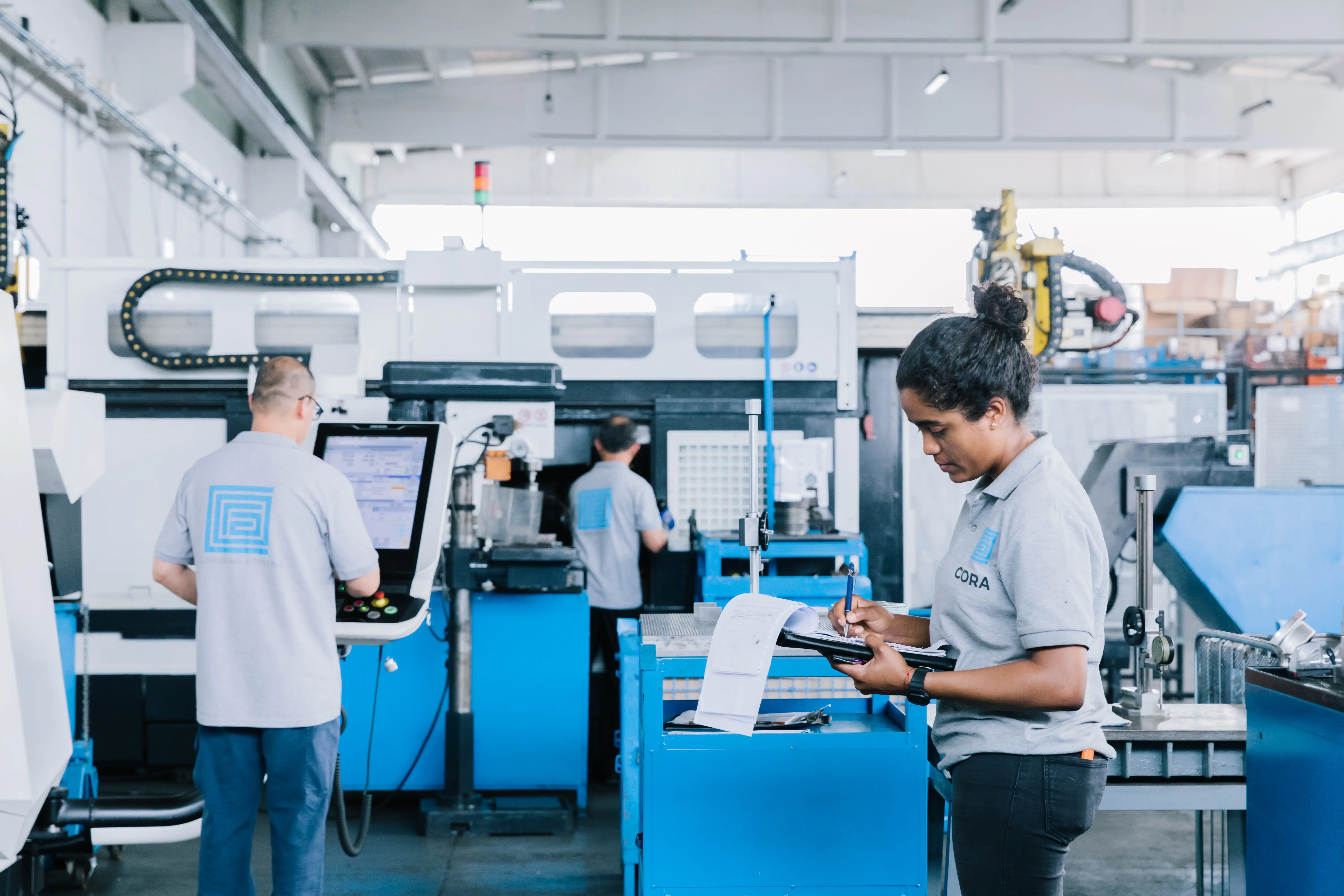 Factory workers in matching gray shirts operating machinery and taking notes on a clipboard.