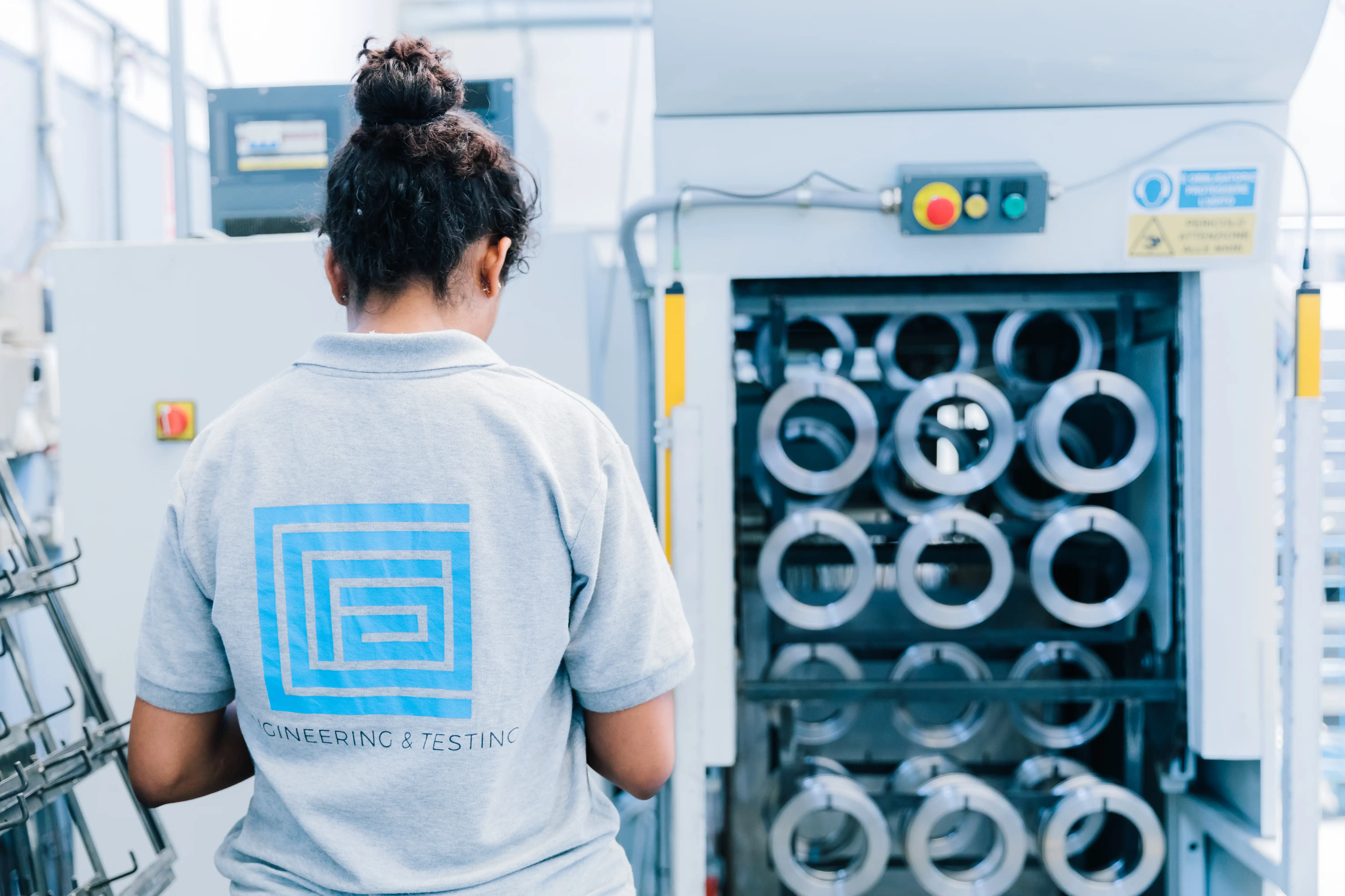 Person in gray shirt with engineering and testing logo stands in front of machinery with circular metal parts.