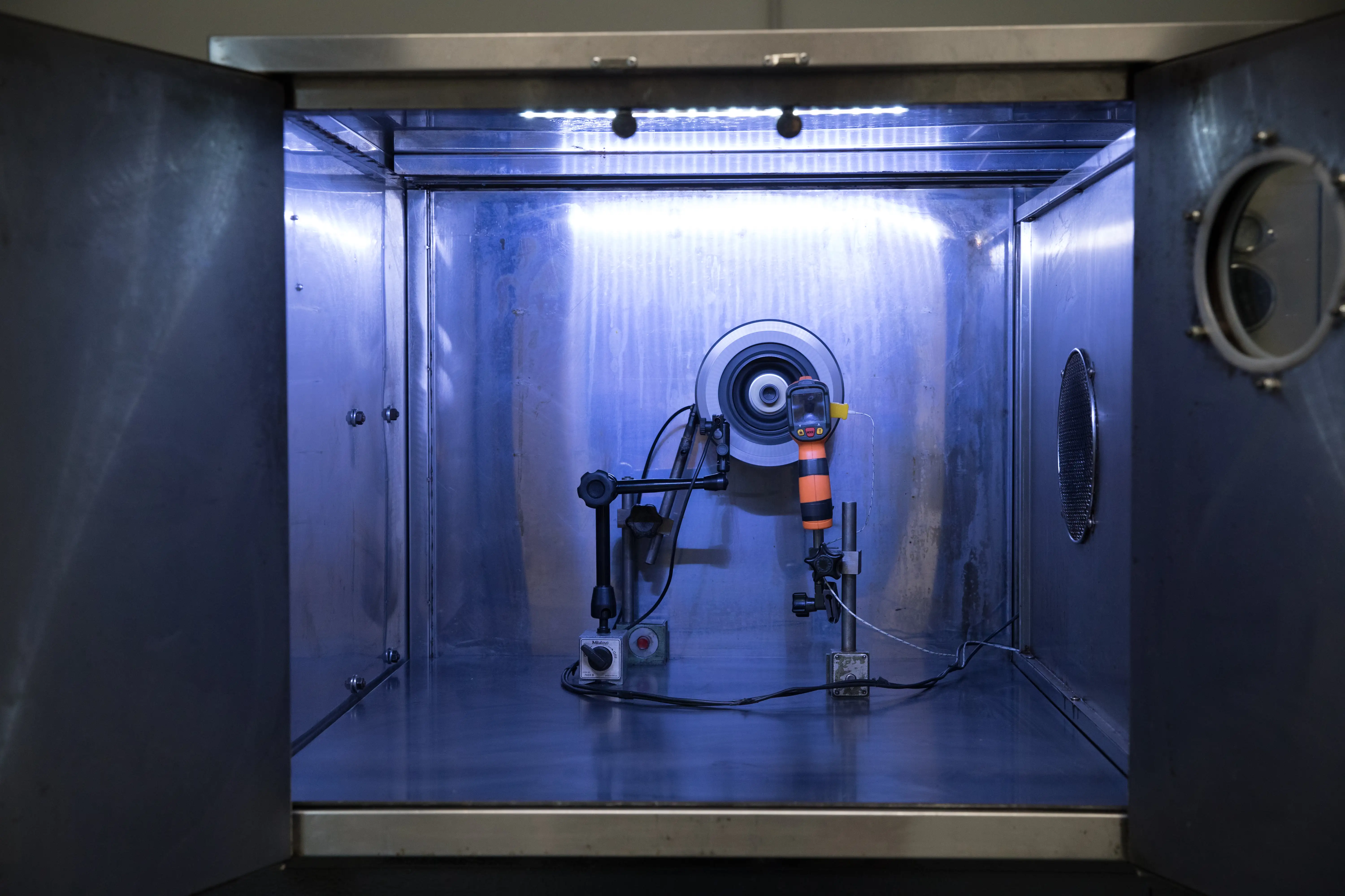 Metallic test chamber with mounted sensor device and connected wires under blue lighting.