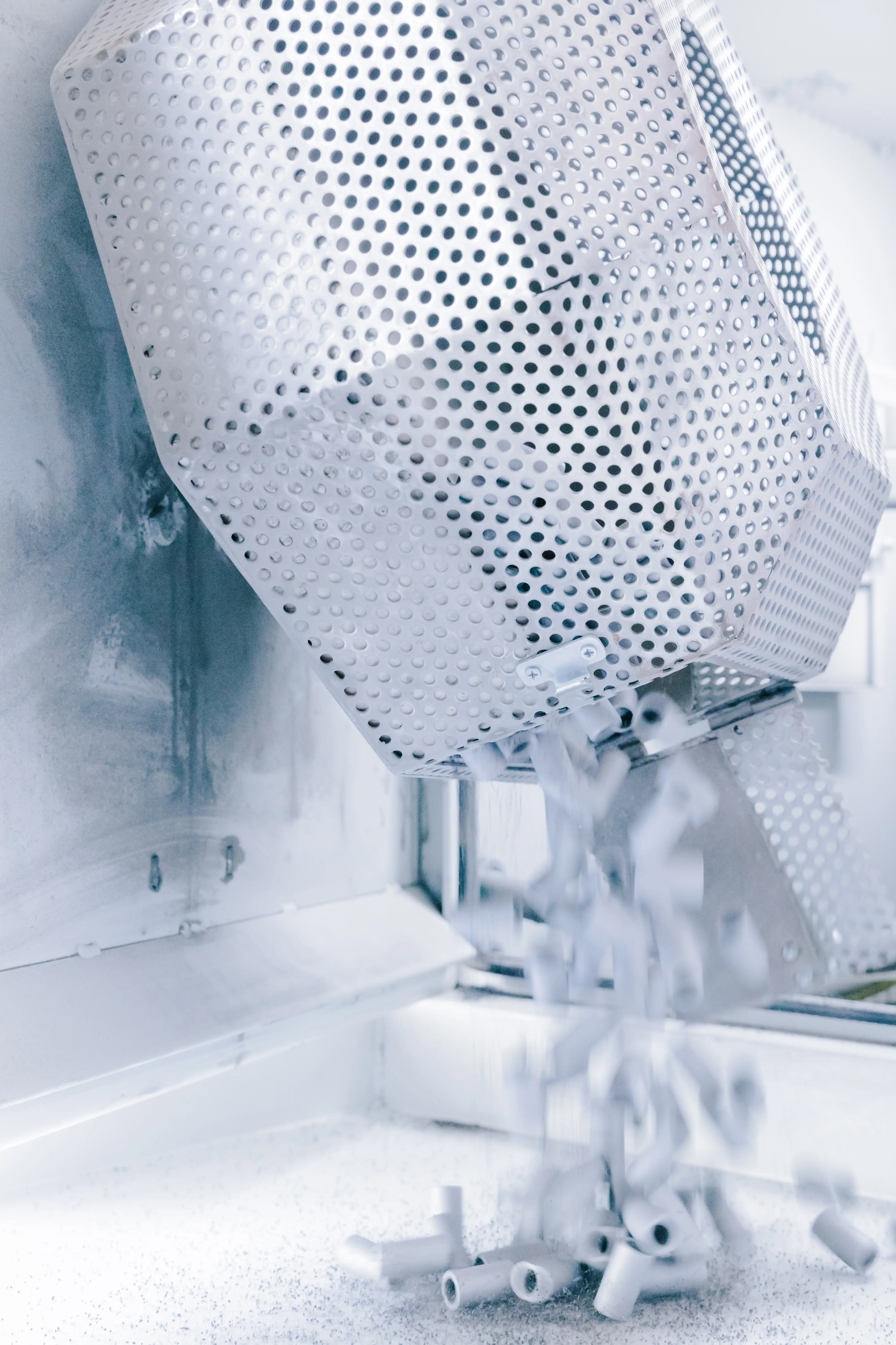 White ceramic pieces falling from a perforated metal container inside a sterile industrial environment.