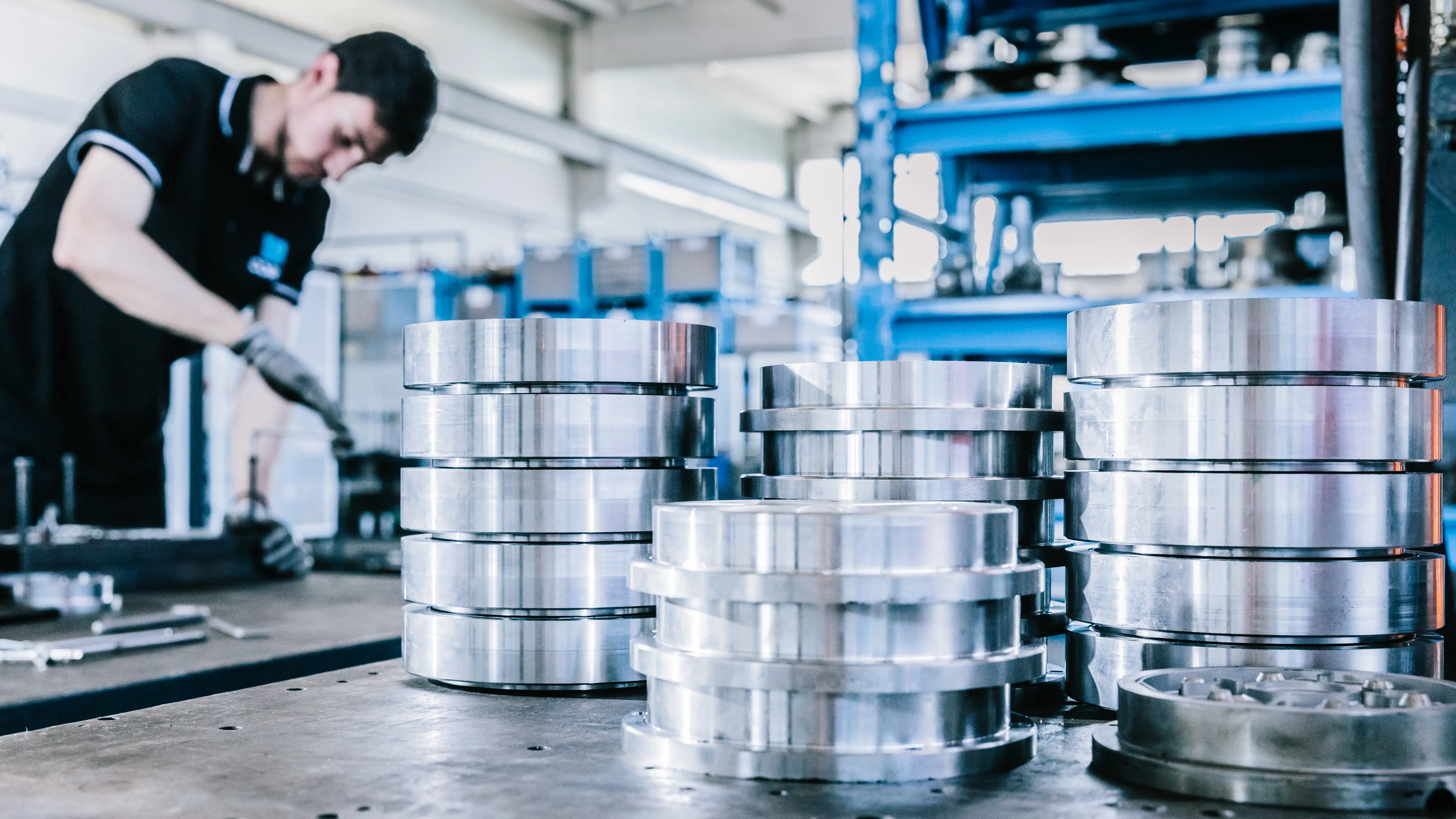 Stacks of shiny metal discs on a workbench with a worker in gloves in the background at a factory.