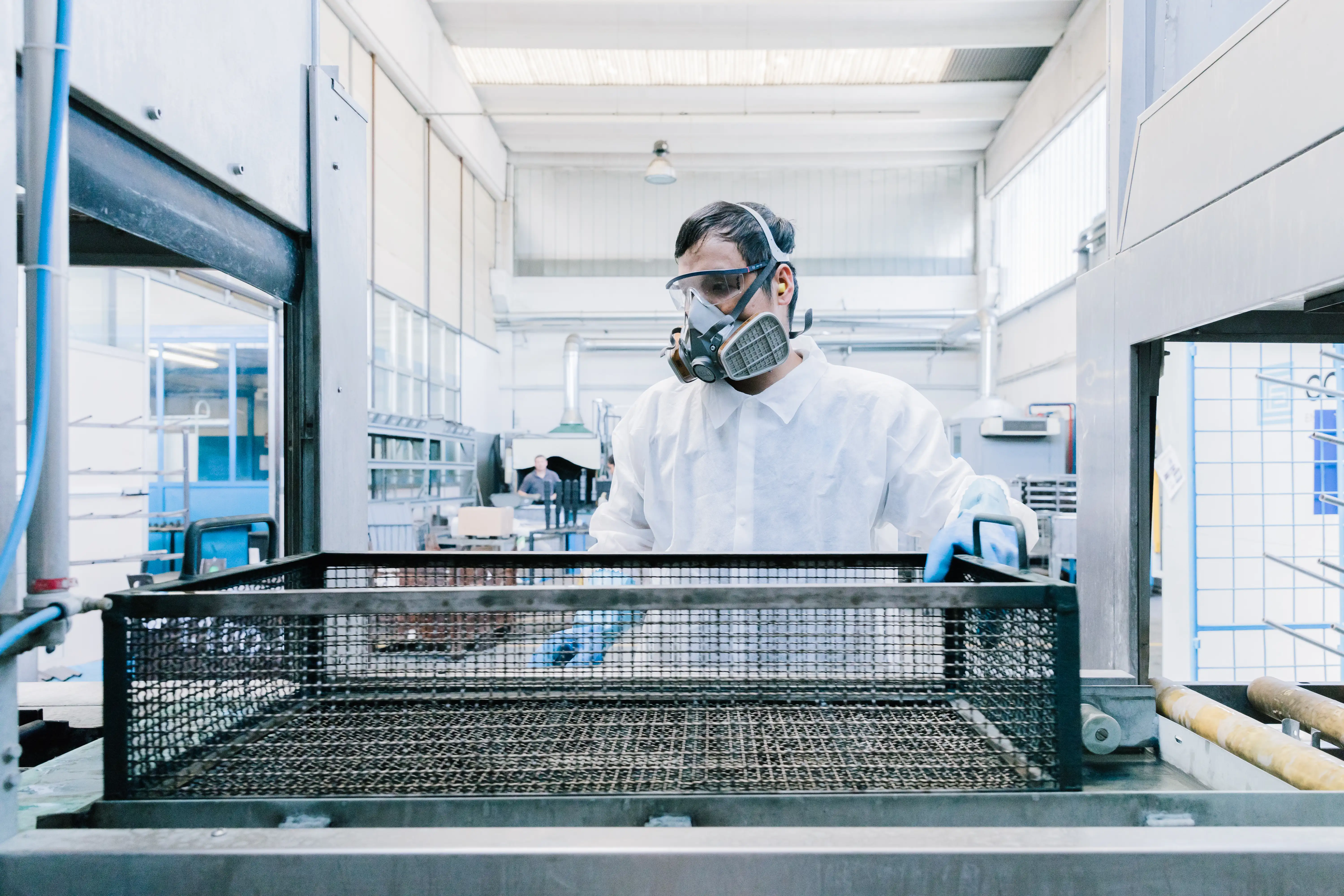 Worker in protective gear operating equipment with a large metal mesh tray in an industrial facility.