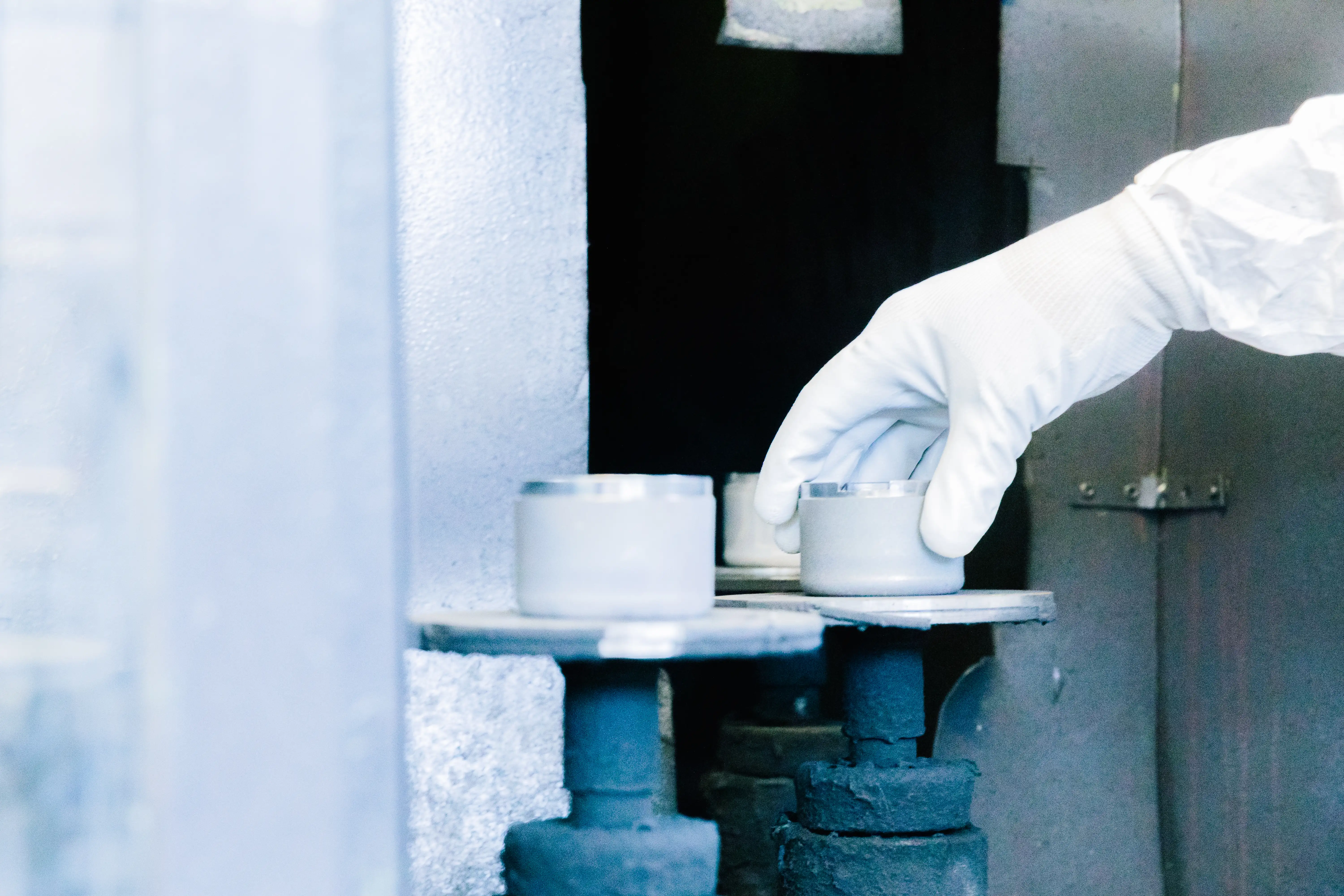 Hand in white glove placing a small container on a metal platform in a lab setting.