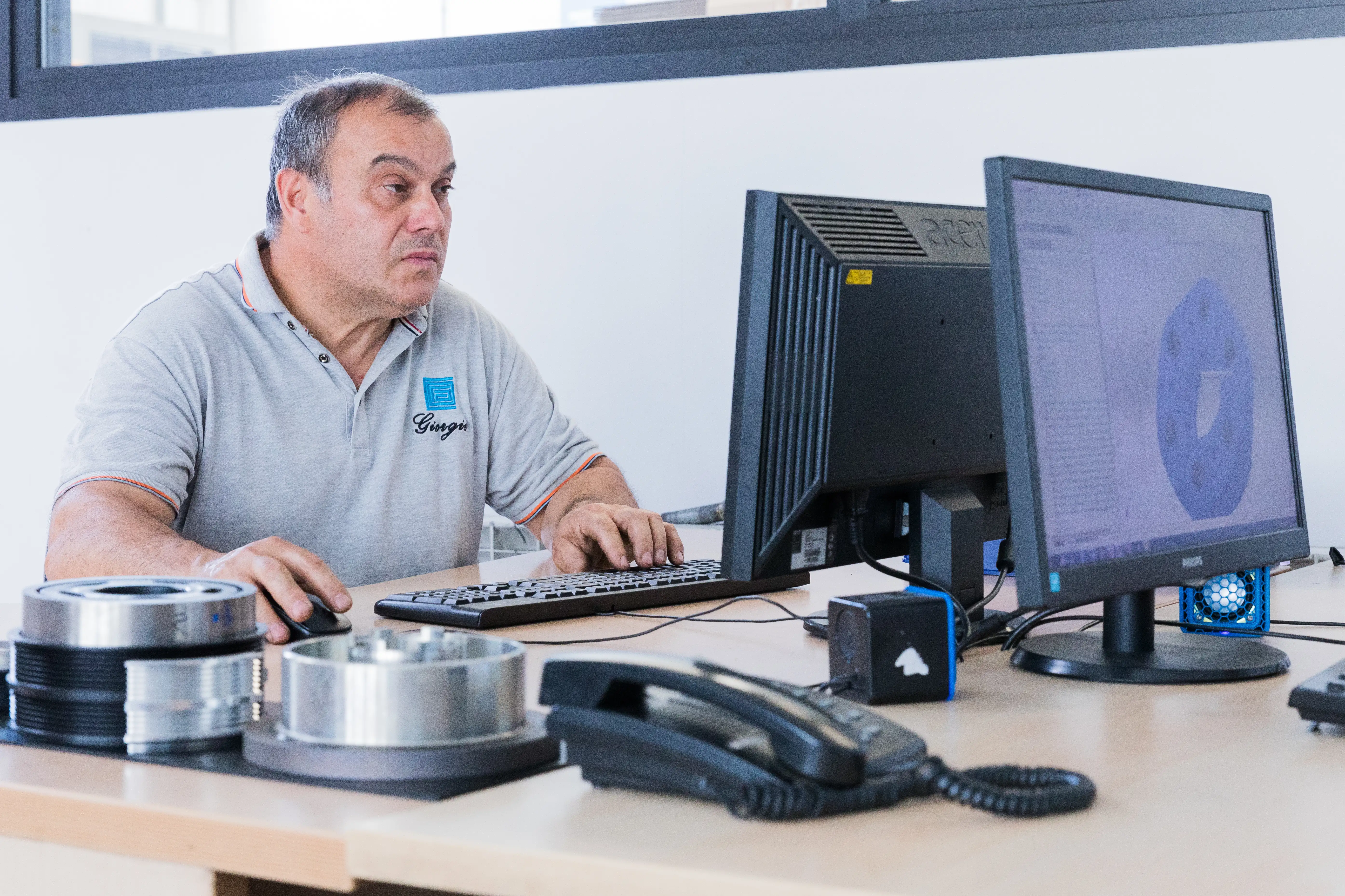 Man in grey polo working on computer at desk with mechanical parts and a telephone.