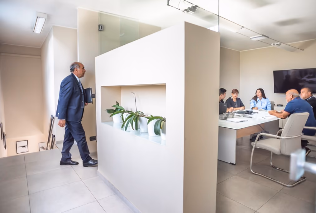 A man in a suit carrying a notebook walking in a modern office corridor with potted plants on a built-in shelf, and a group of five people having a meeting around a table in a conference room.