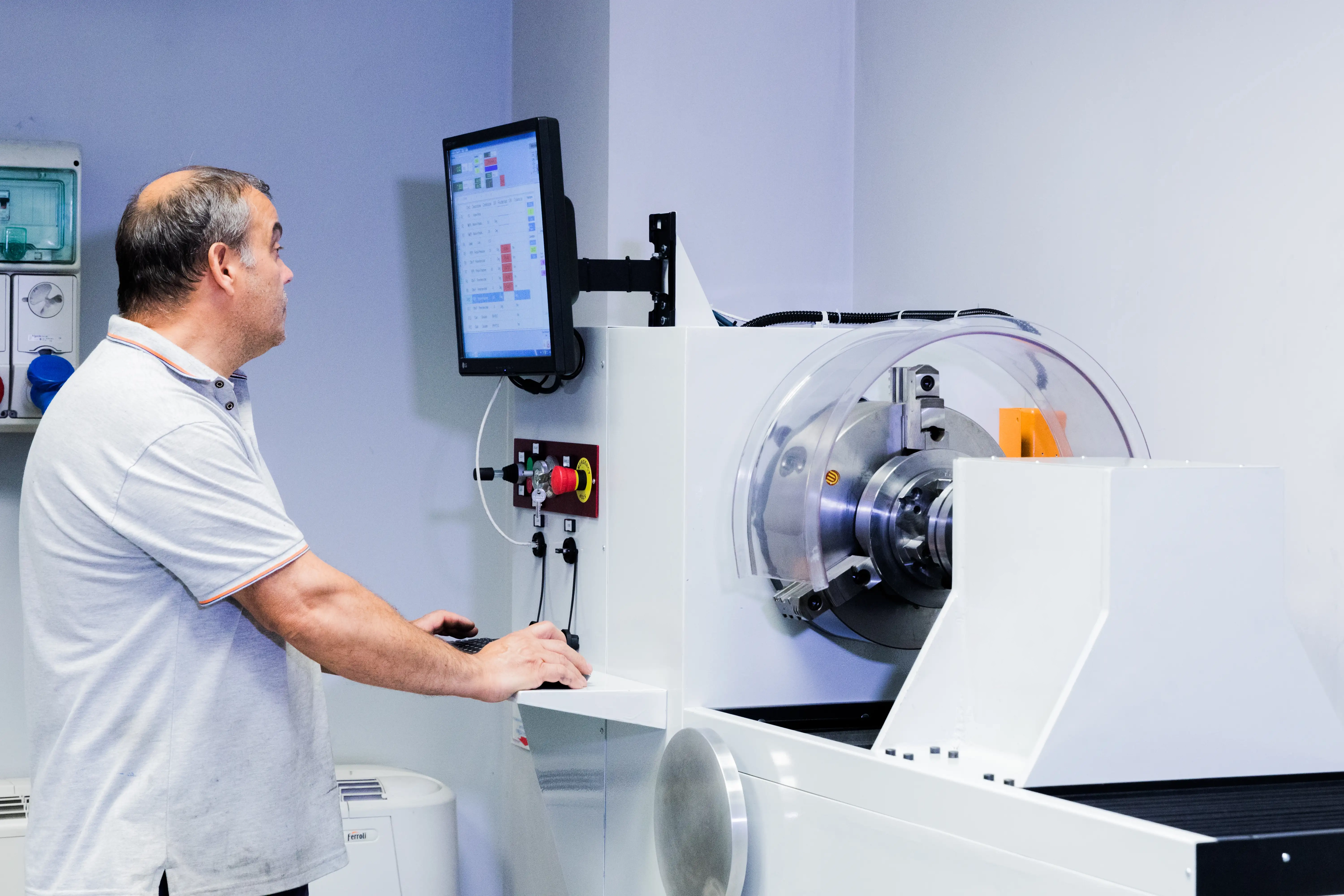Man in a light gray polo shirt operating a white industrial machine with a circular component and a mounted screen displaying data in a room with white walls.