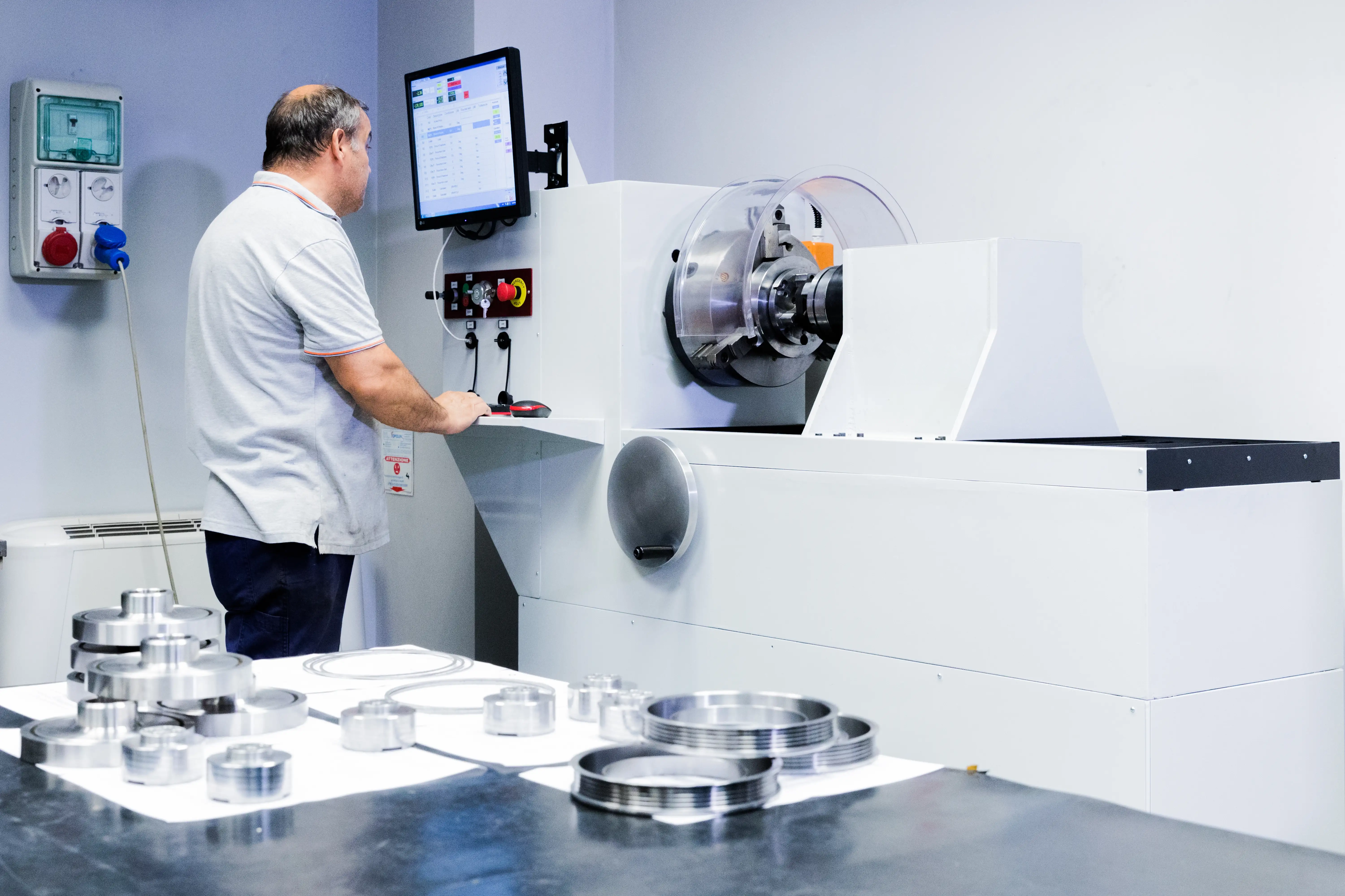 Man operating a white industrial CNC machine with a control panel and monitor, metal parts on a table in the foreground.