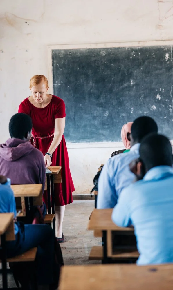 Teacher in red dress assisting students seated at wooden desks in a classroom with a blackboard.