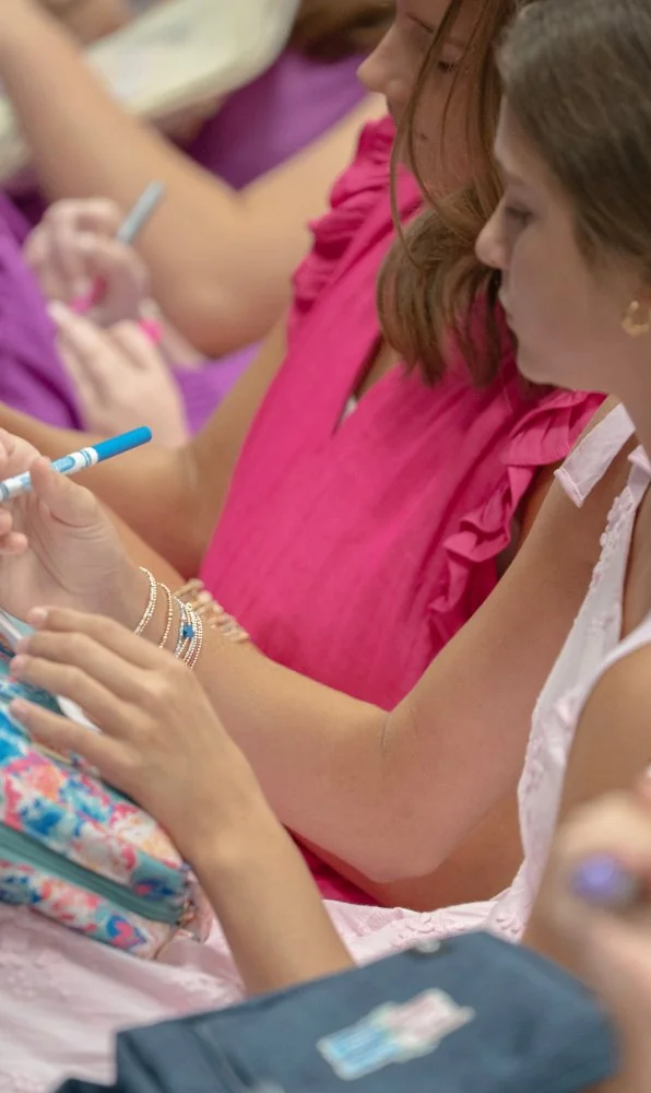 Teen girls sitting closely together, writing notes during a study session, with focus on their hands and pens.