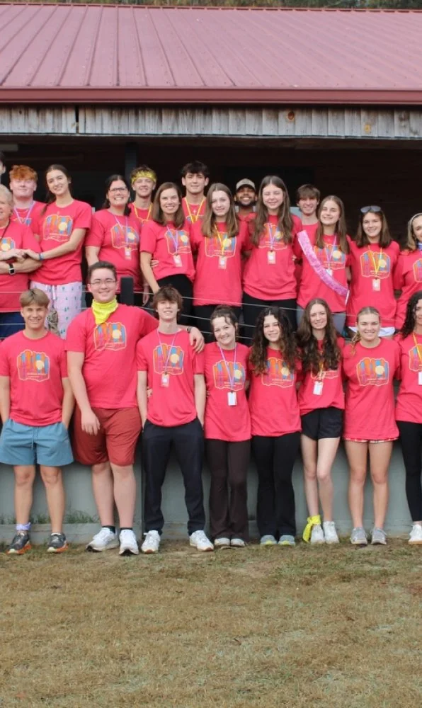 Group photo of smiling youth wearing matching red camp t-shirts standing outdoors in front of a building with a red metal roof.
