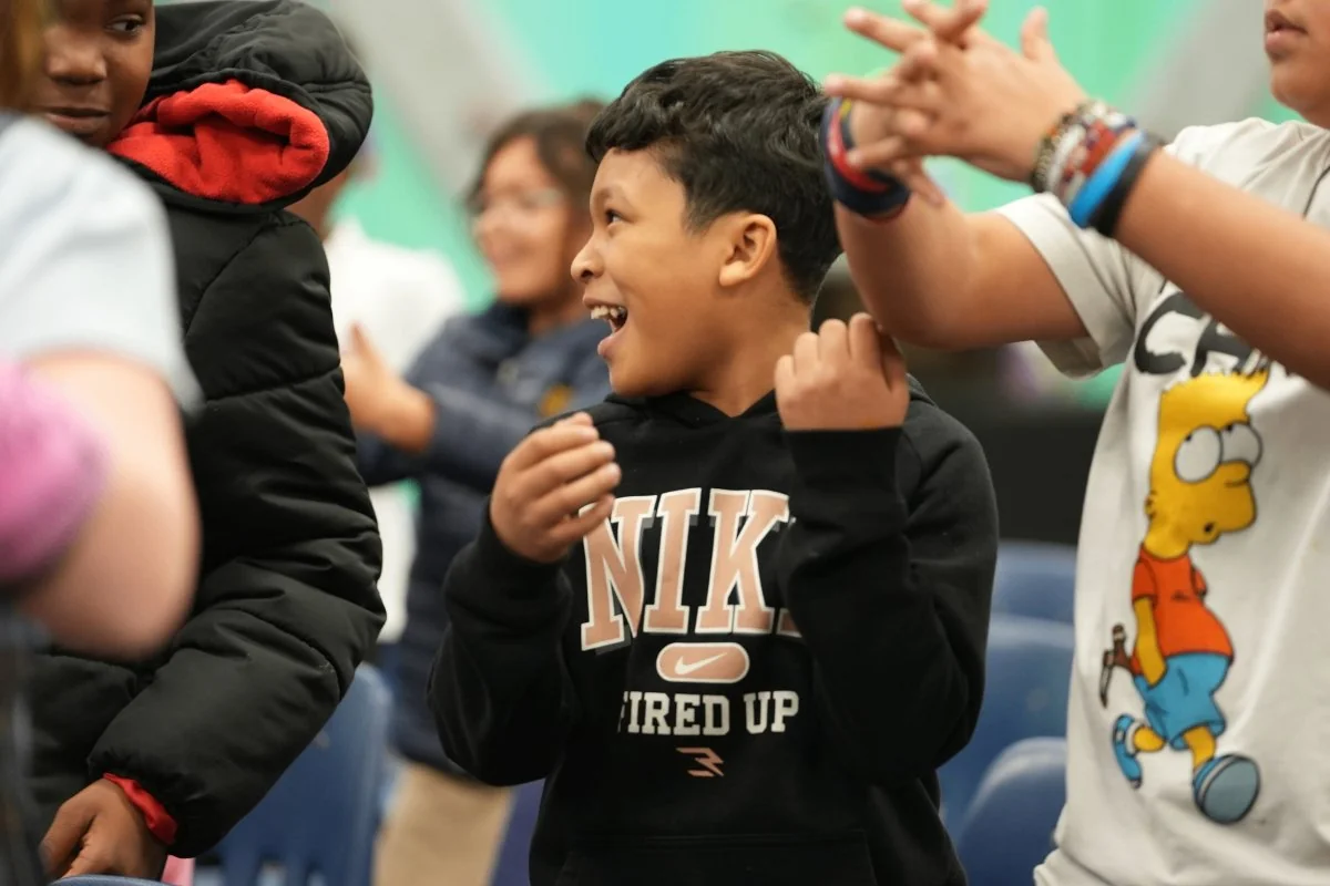 Smiling boy in a black Nike hoodie clapping and looking to his left during a group event.