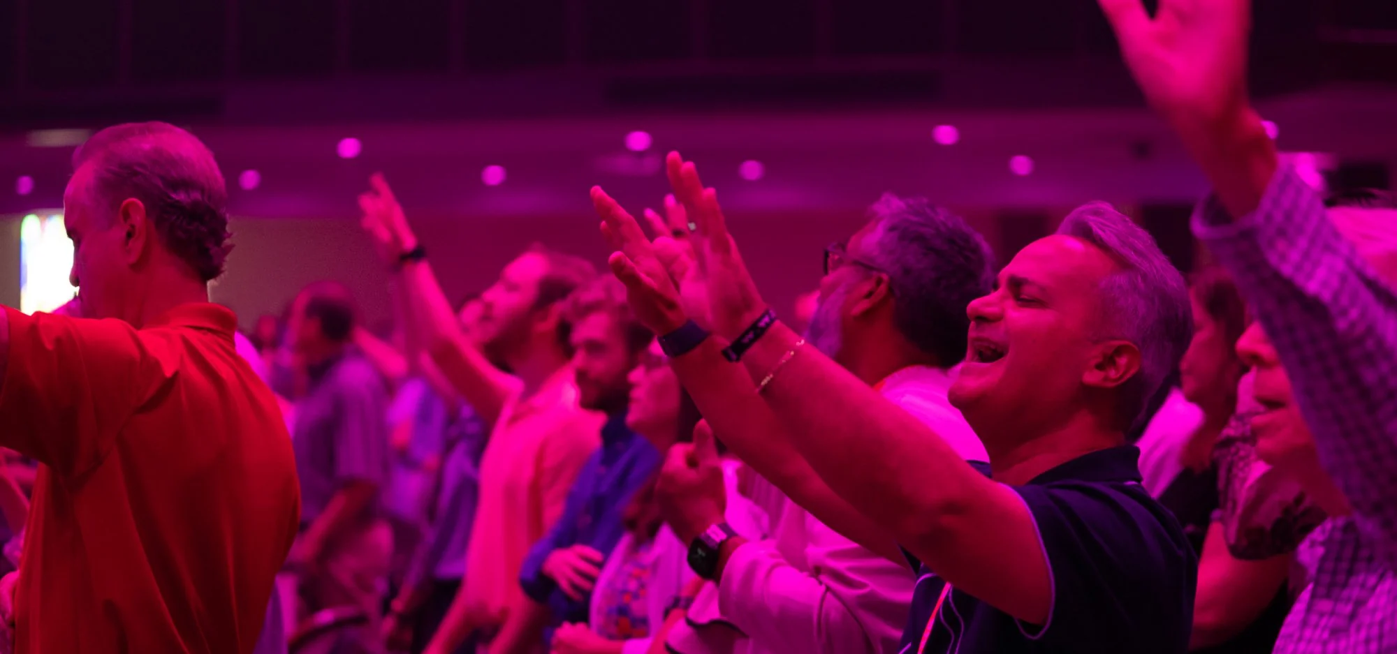 Congregation with raised hands worshiping under purple stage lighting.