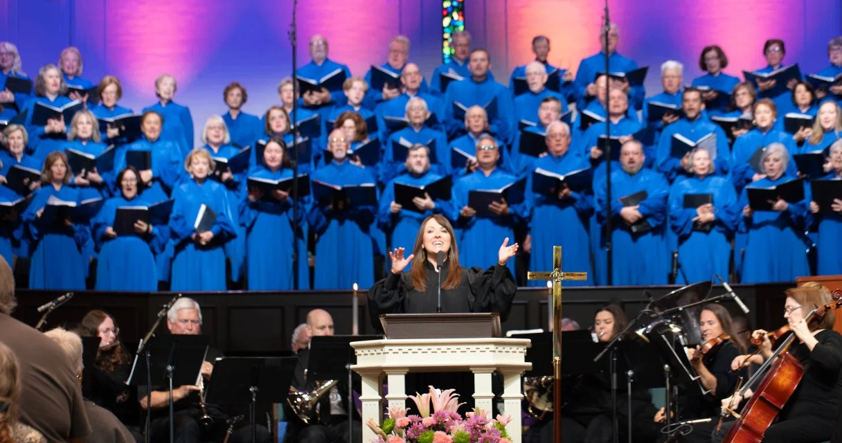Woman preacher speaking at a white pulpit with a choir in blue robes and musicians playing instruments behind her.