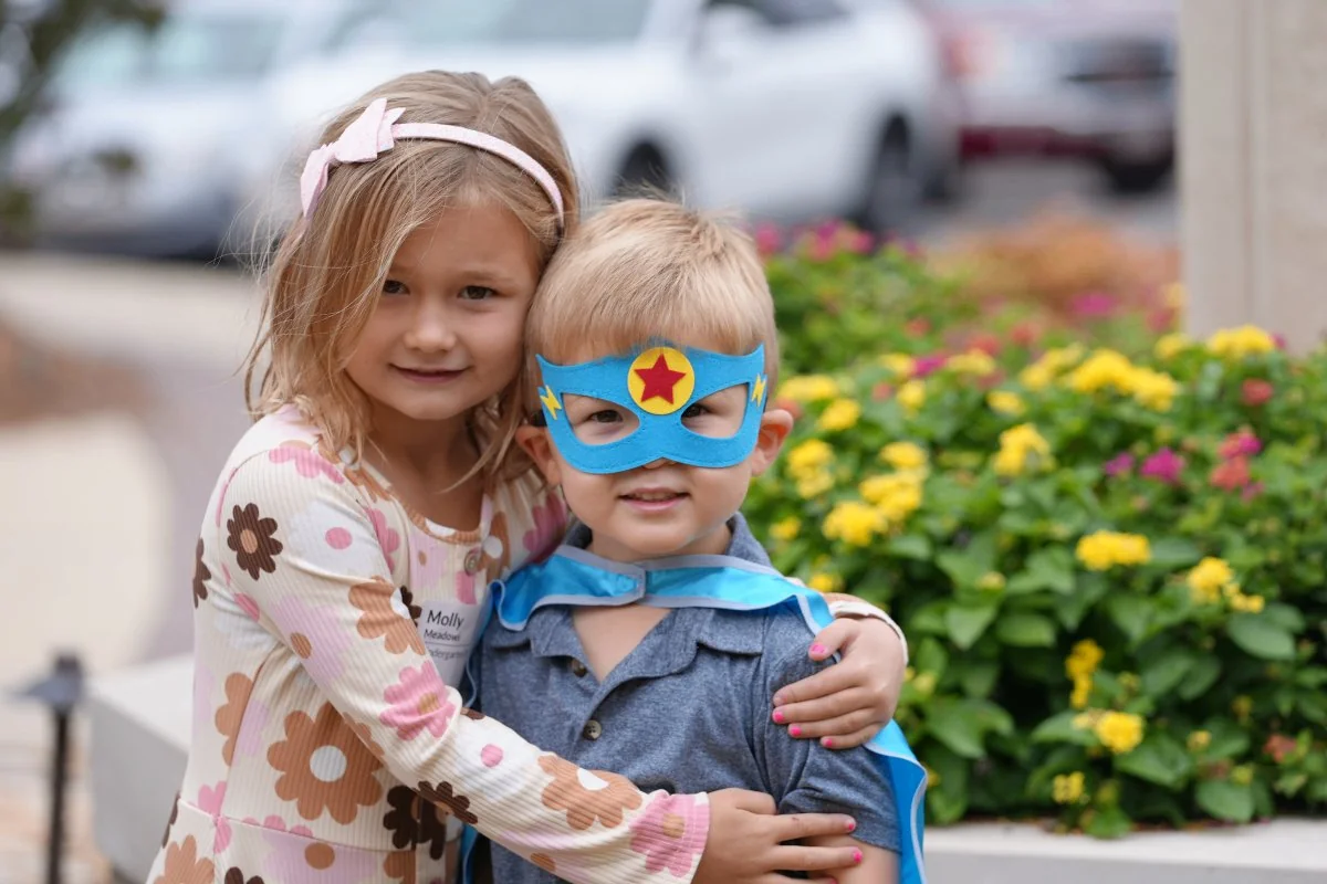 Young girl with floral dress hugging a boy wearing a blue superhero mask and cape outdoors near flowers.