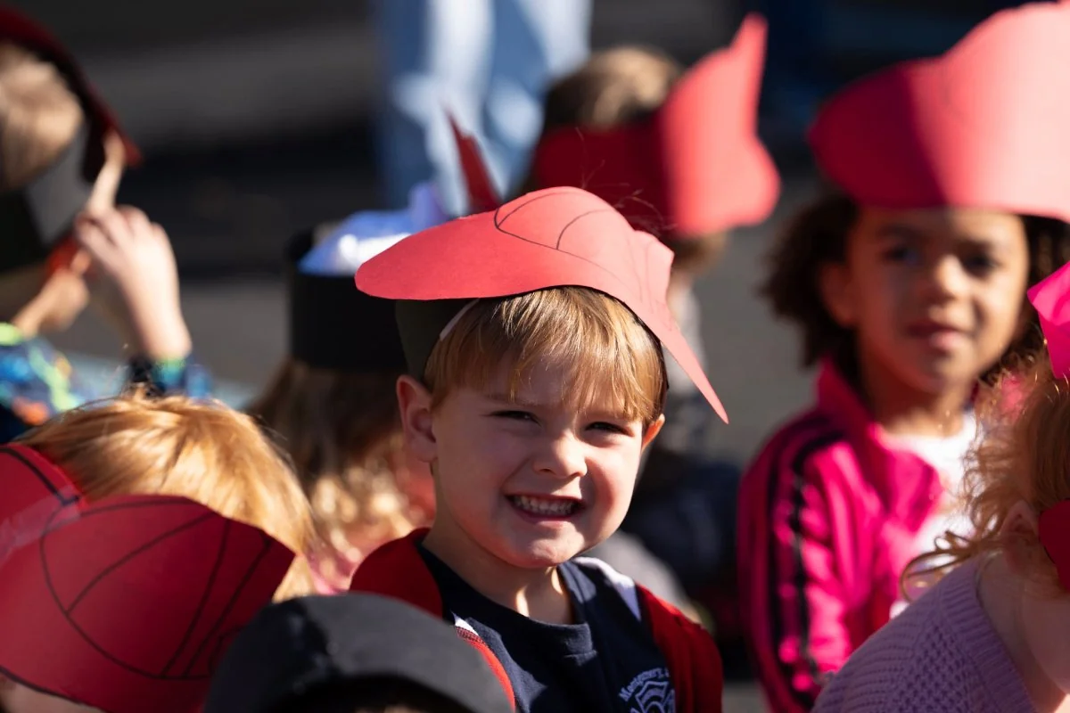Smiling young boy wearing a red paper firefighter hat among other children also wearing similar hats.