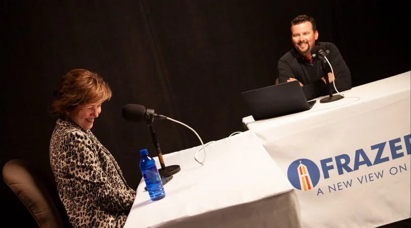 Two podcast hosts sitting at a table with microphones and a laptop, smiling during a recording session with a Frazer banner on the table.