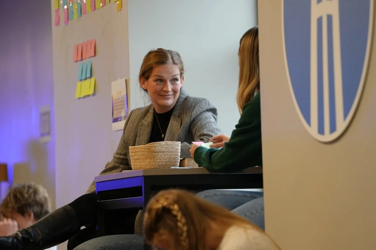 A woman in a gray blazer sits at a table, engaging in conversation with another person in a green sweater, with colorful sticky notes on the wall behind them.