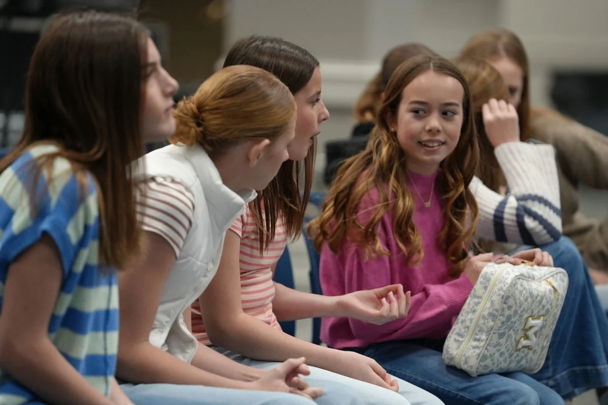 Group of middle school girls sitting and talking indoors, one girl in a pink sweater holding a white patterned bag.