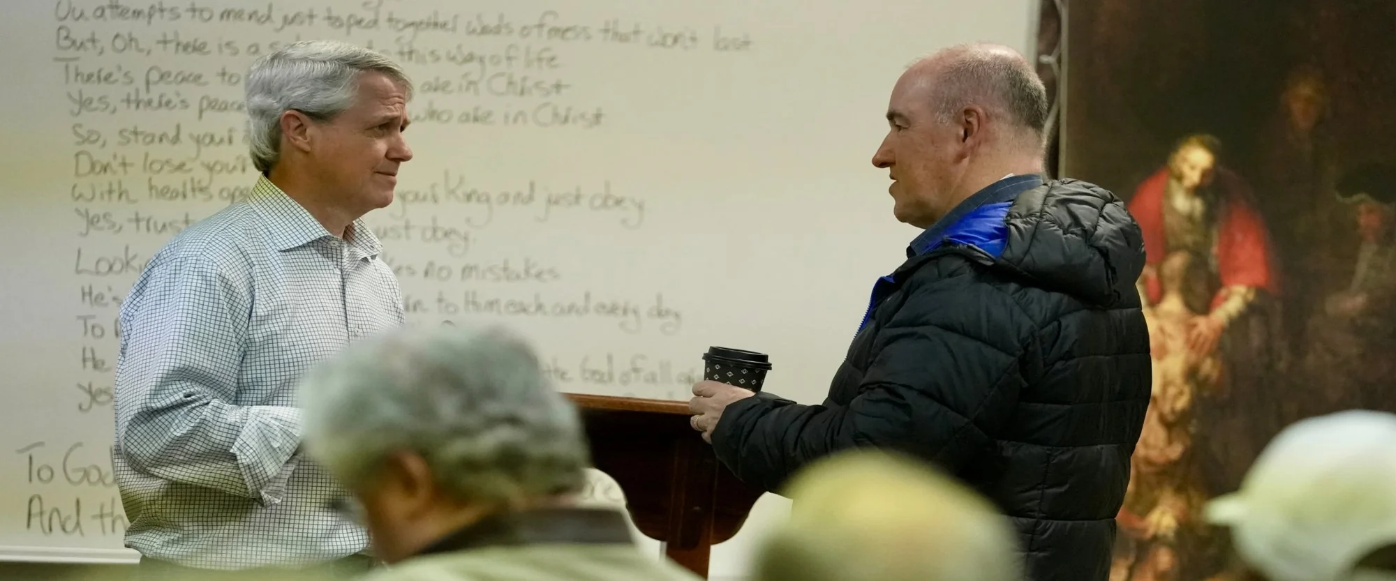 Two men engaged in conversation in a classroom setting with a whiteboard and religious painting in the background.