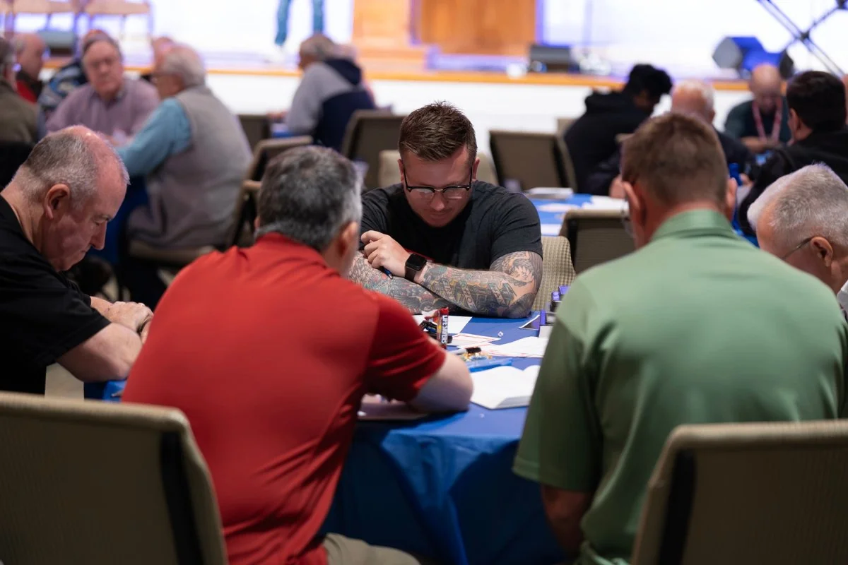 Group of men sitting around a table with blue tablecloth engaged in a Bible study or discussion, with one man in glasses and tattoos focused on reading.