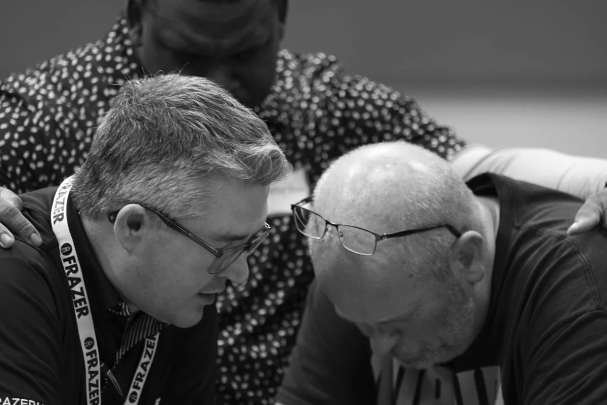 Three men with heads bowed in close prayer, one wearing a lanyard with the name Frazer.