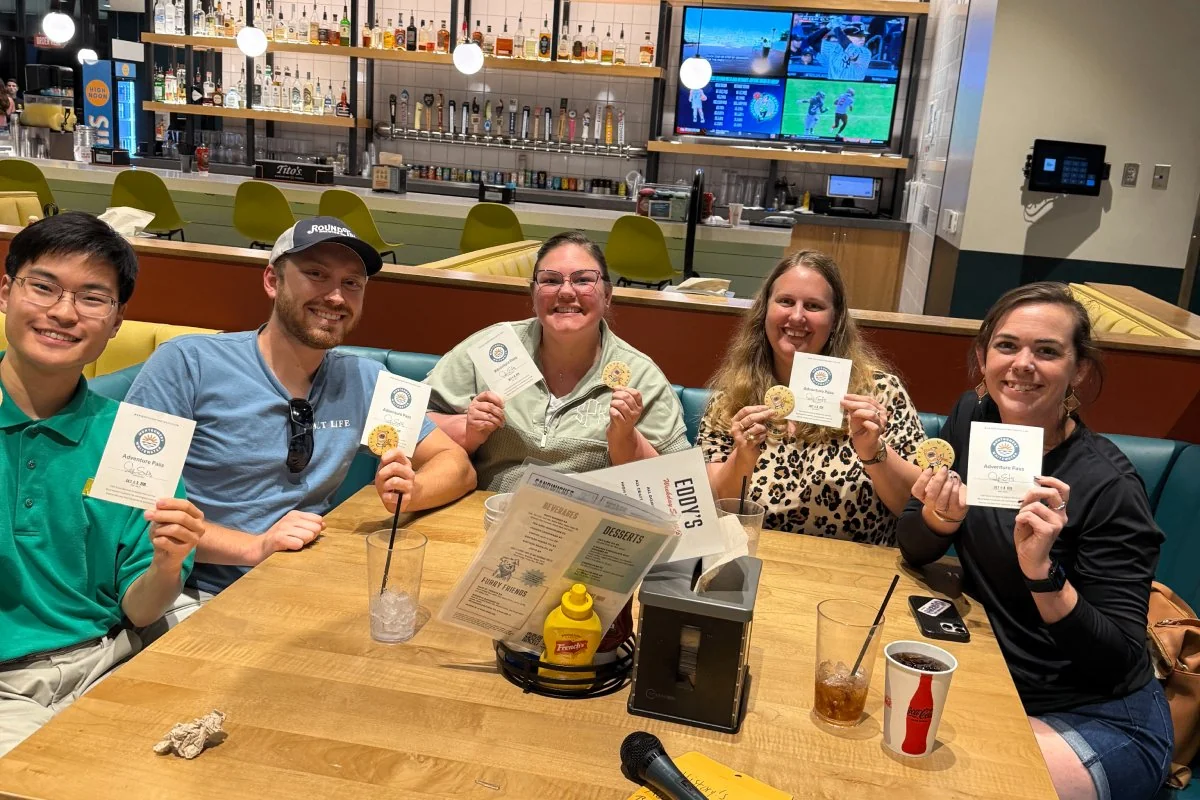 Five smiling people sitting at a restaurant table holding certificates and cookies, with drinks and menus on the table.