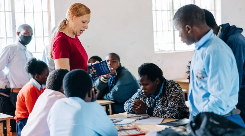 A woman in a red shirt reading from a book to a group of students seated around tables in a classroom.