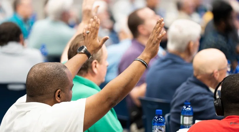 Man raising both hands in worship during a conference with other attendees seated around him.