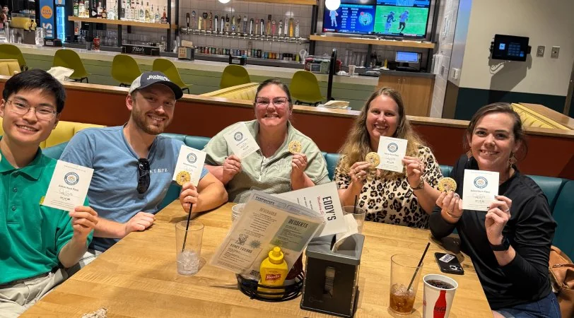 Five friends sitting at a restaurant table smiling and holding cookies with paper cards.