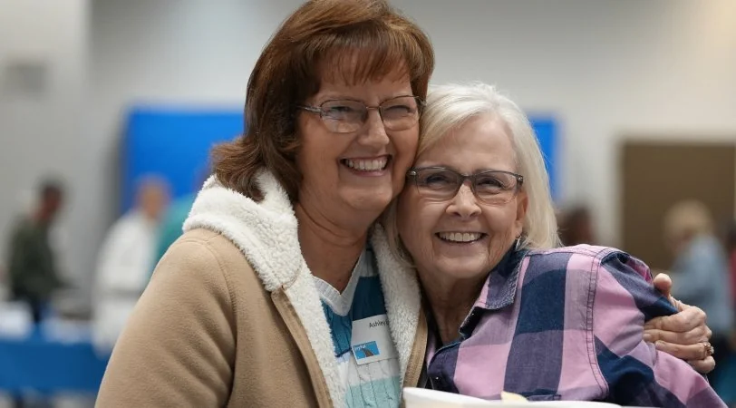 Two middle-aged women wearing glasses smiling and hugging indoors, one in a tan coat and the other in a plaid shirt.