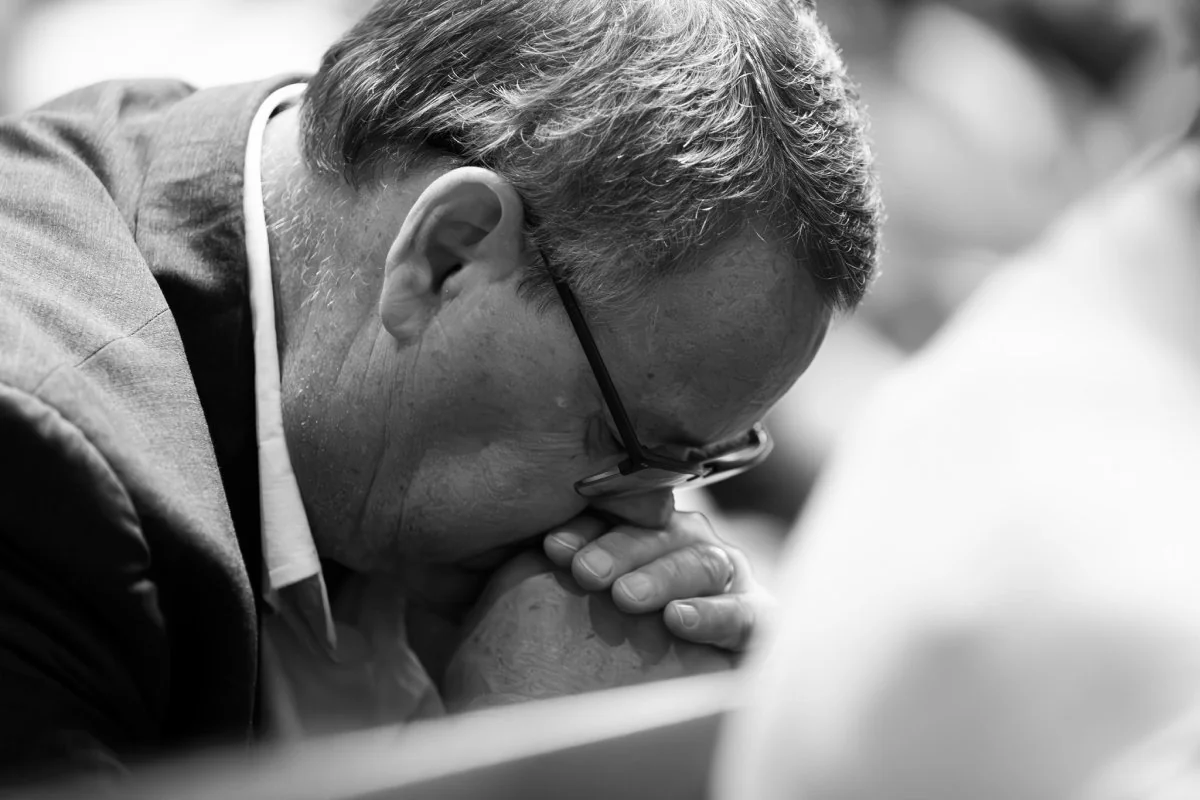 Black and white close-up of a man with glasses bowing his head and clasping his hands in prayer.