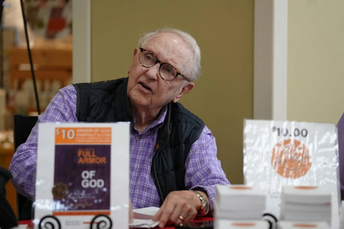 Elderly man with glasses and a black vest sitting at a table signing books titled 'Full Armor of God' priced at $10.