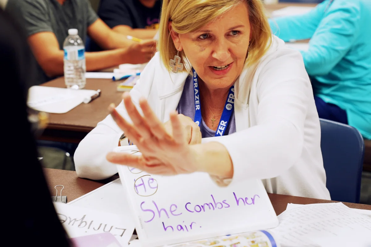 Woman teacher showing a whiteboard with the sentence 'She combs her hair.' in a classroom setting.